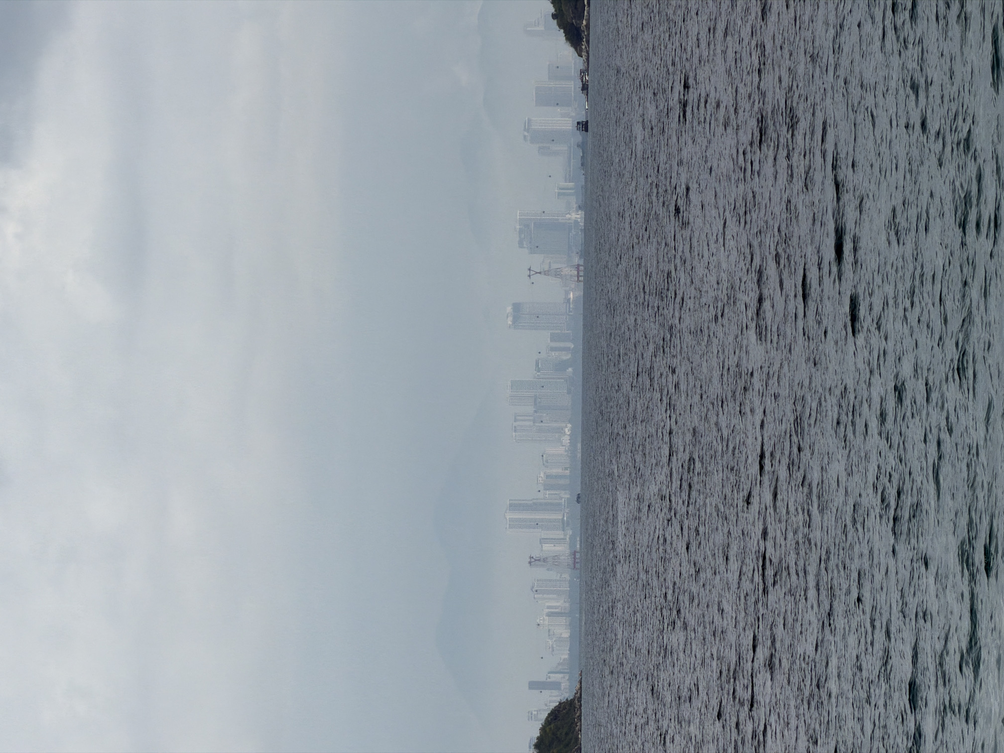 The image depicts a cityscape viewed across water, likely taken in Nha Trang Ward, Vietnam. The right side of the image is dominated by choppy grey water. In the background, a cityscape is visible through a hazy atmosphere. The buildings appear to be a mix of high-rise and mid-rise structures. The sky above is overcast, further contributing to the hazy and muted color palette of the scene. Vegetation is visible on the immediate left and right edges of the water, suggesting a shoreline perspective.