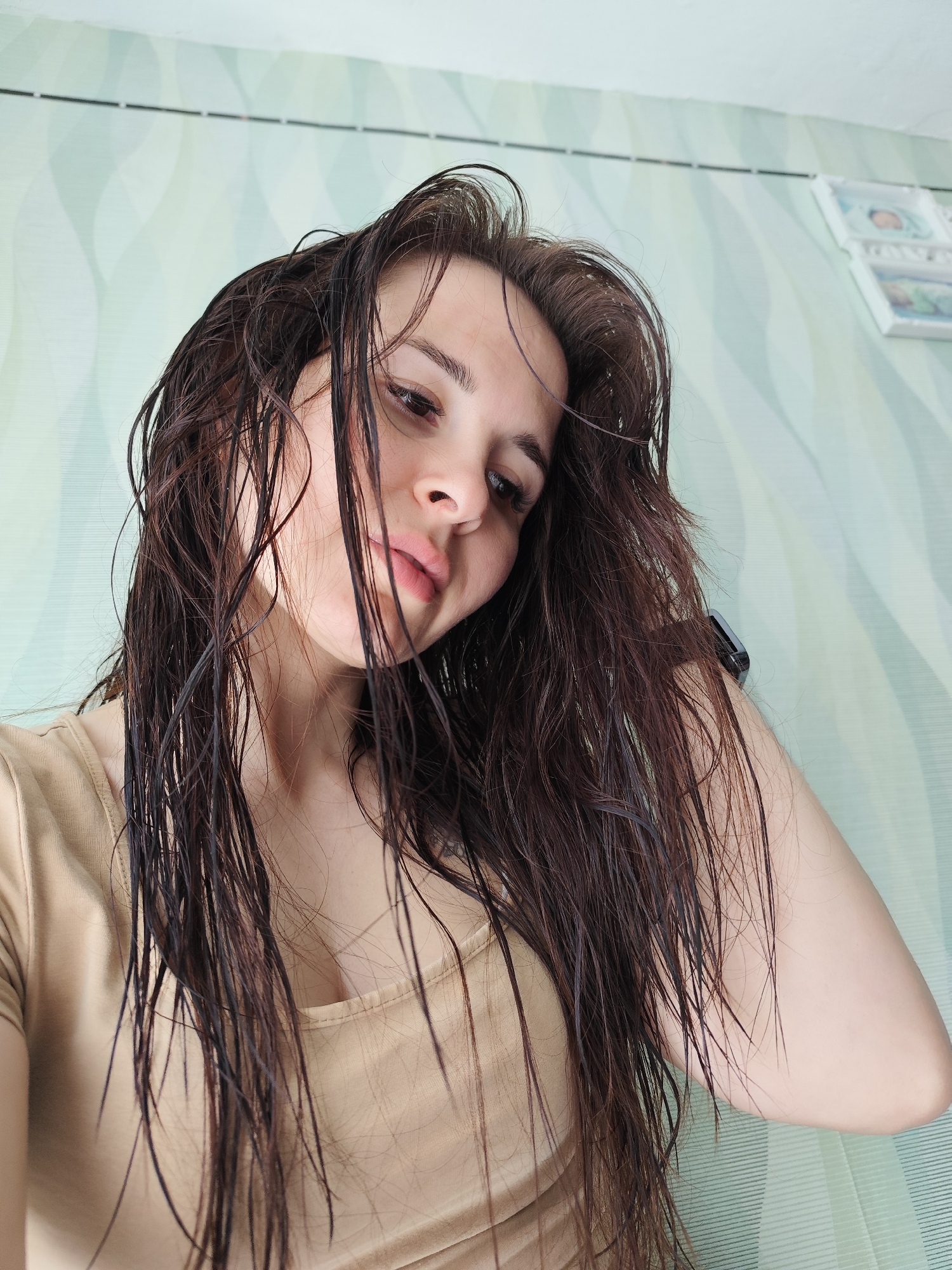 A selfie features a young woman with long, dark brown hair that appears wet or heavily styled, hanging in defined strands around her face and shoulders. She is looking downwards towards the camera with a neutral to slightly soft expression, her lips slightly parted. She wears a plain, light beige or nude-colored top with a rounded neckline.
The background consists of an indoor wall covered in light green wallpaper with a subtle, wavy pattern. Towards the upper right, two small, white-framed items, possibly pictures or photographs, are mounted vertically on the wall. A white ceiling is visible at the top edge of the image. On the woman's right wrist, a dark-colored object, resembling a smartwatch or fitness tracker, is visible. The lighting is bright and even, suggesting an indoor daytime setting. The scene conveys a personal and casual moment, possibly after washing her hair. The location context for this image is Фоки, Russia.