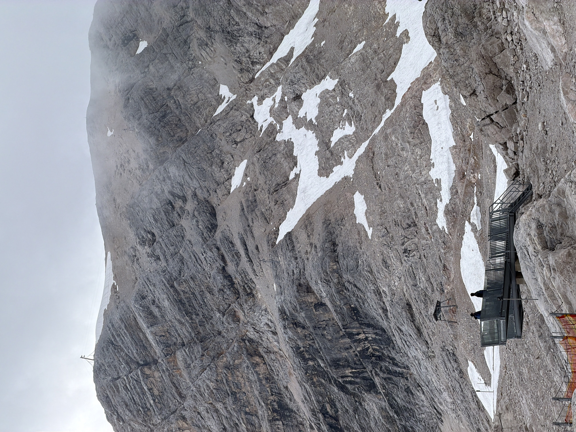 The image shows a rugged, rocky mountain landscape with patches of snow. In the foreground, there is a metal viewing platform with two people standing on it. The platform is elevated and has a railing. Behind the platform, a small, dark structure with a roof is visible on the mountainside. The mountain is steep and heavily striated with rock formations. The sky is overcast and appears hazy. The location is likely the Zugspitze, Germany's highest peak, near Garmisch-Partenkirchen.