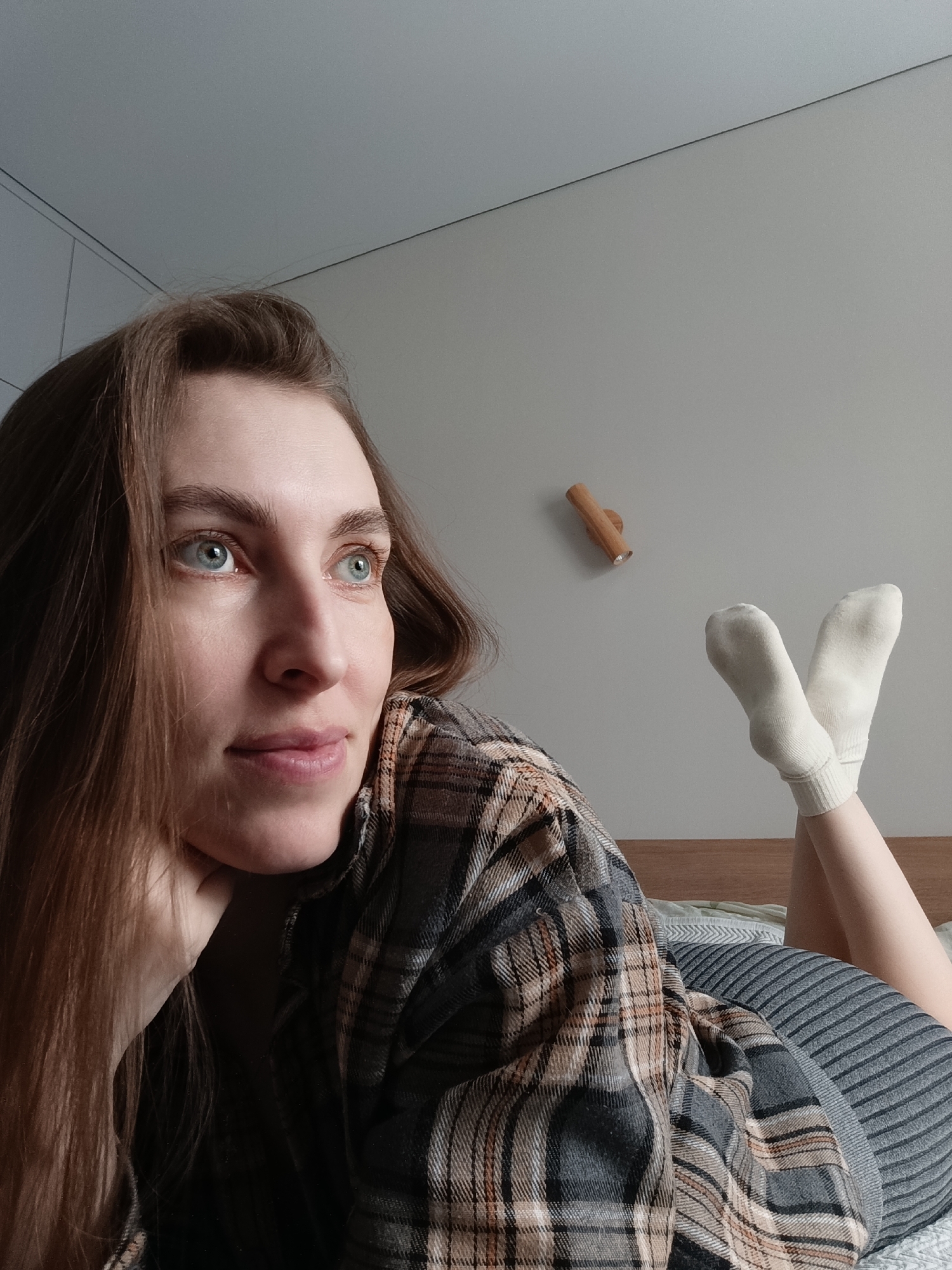 A woman lies on a bed, resting her head on her hand, with her feet up in the air wearing white socks. She is wearing a flannel shirt and a striped garment. The background consists of a wall with a wooden object affixed to it. The scene is likely indoors in a home in Russia. There is insufficient information to confirm or deny the location is Chaikovsky, Russia.