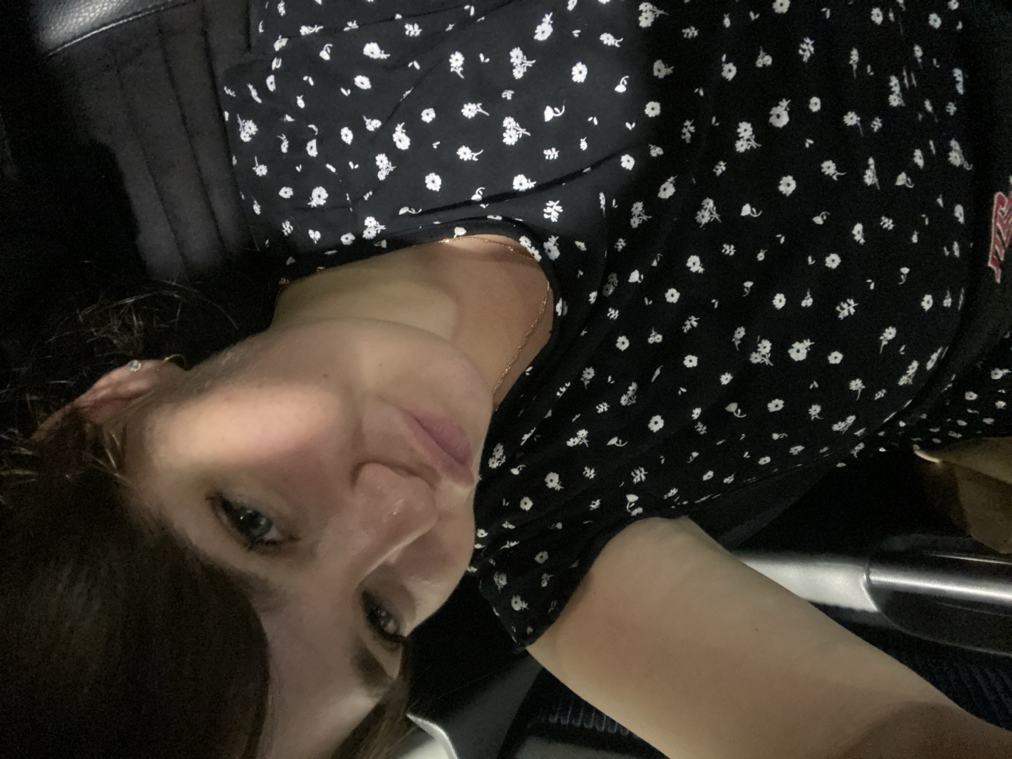 In the city of Italmas, Russia, a young woman with dark hair, appearing to be in her early twenties, is captured in a close-up selfie. She is seated, possibly in a vehicle, given the dark upholstery visible behind her. The woman has fair skin, blue eyes, and is wearing a black dress with a white floral pattern. Her hair is pulled back, revealing a small earring in her left ear. She is looking directly at the camera with her lips slightly pursed, creating a playful expression. A thin gold necklace is visible around her neck. The lighting is dim, suggesting an indoor or evening setting. The background is dark and out of focus, emphasizing the subject.