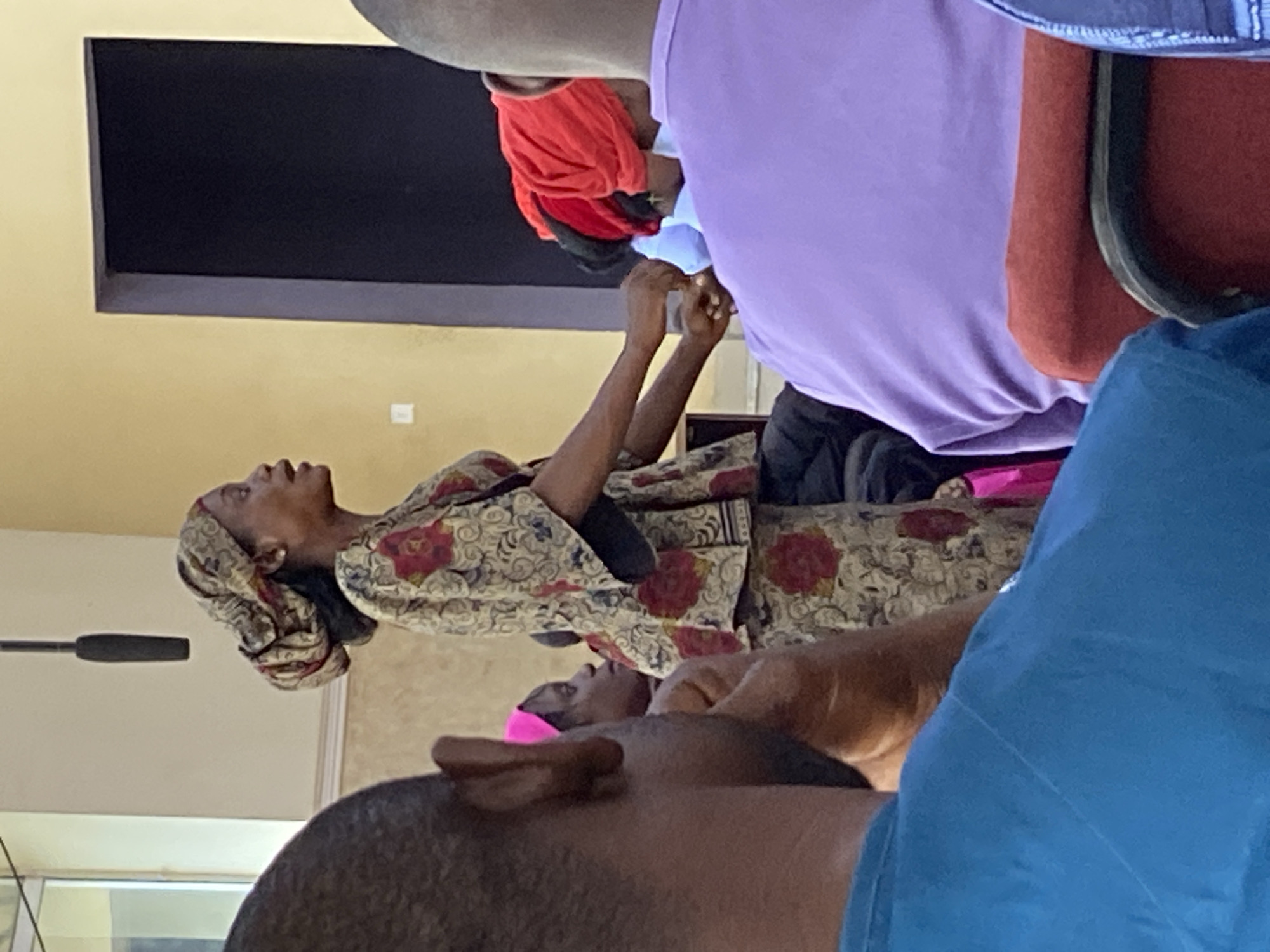 The image shows an indoor scene in Mararaba, Nigeria. A woman wearing a patterned headscarf and a matching floral dress is seated, looking upwards with her hands raised and clasped together. In front of her, another person with a pink headscarf is seated, and the head and shoulders of a man in a blue shirt are visible. To the woman's right, there are people with red and purple shirts, indicating that there is a group of people sitting together. There is a dark rectangle on the wall, likely a television or monitor, and the walls are painted yellow. A microphone is also visible.