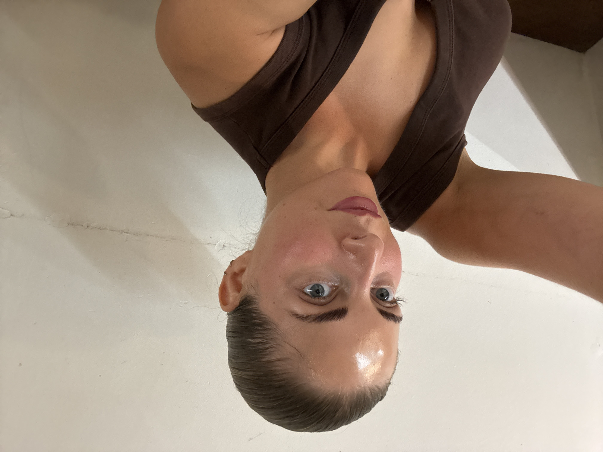 A woman is taking a selfie against a white wall. She has dark hair pulled back, blue eyes, and is wearing a brown tank top. Her left arm is extended towards the camera, and her right shoulder and neck are visible. The background is a plain white wall with some minor imperfections. The scene does not appear to be in Paris, France.