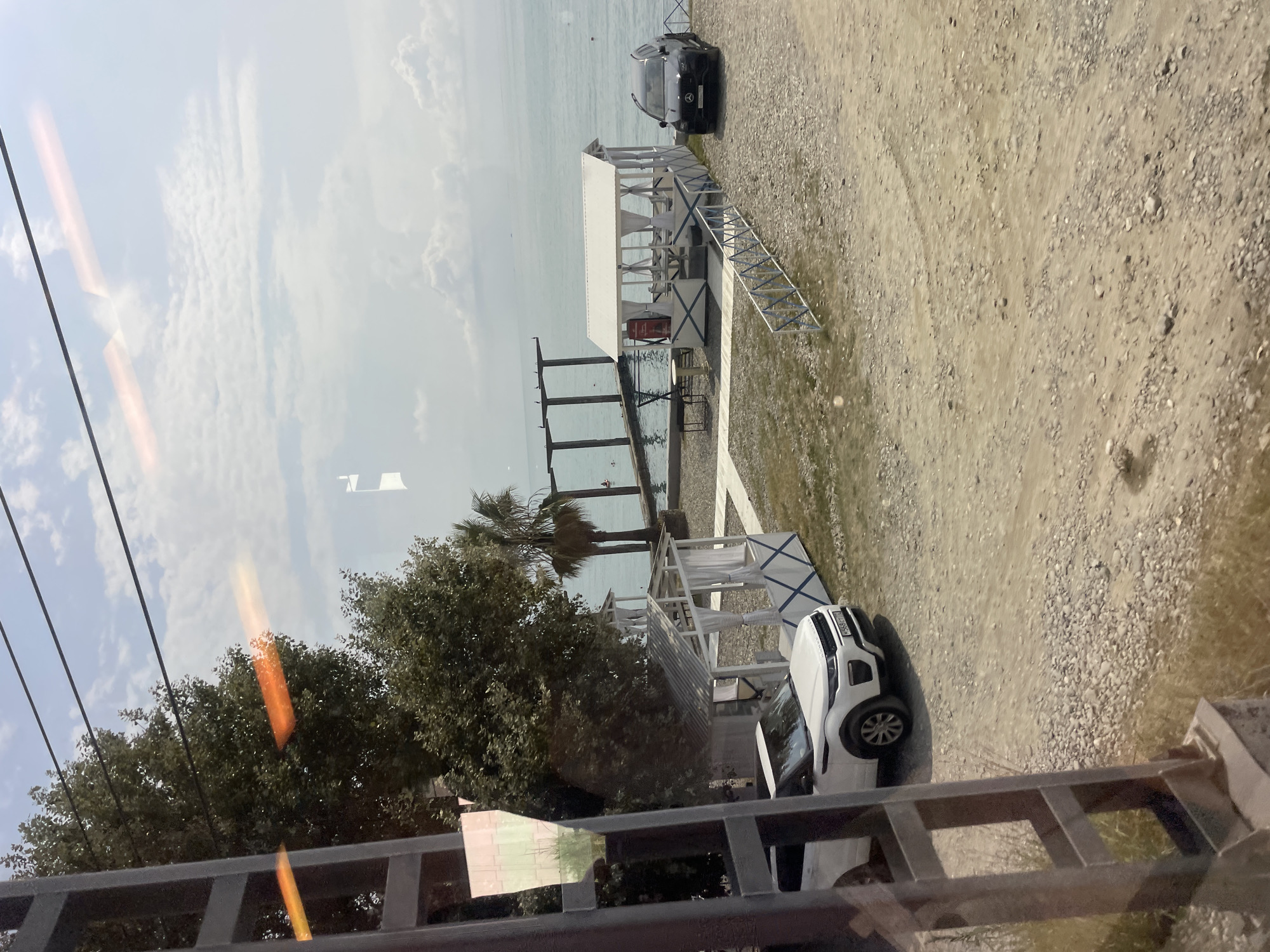 A clear, bright, daytime, eye-level shot captures a scene on a rocky, sparsely vegetated beach area near the sea in Sochi, Russia. The foreground is dominated by a gravelly path and a metal structure, possibly part of a fence or support. Two cars are parked on the beach: a white SUV on the left and a black SUV on the right, positioned near a white, open-sided gazebo with a pitched roof, featuring white curtains and blue decorative trim. Several tables and chairs are visible inside the gazebo.
Beyond the beach, the calm, turquoise sea stretches to the horizon, meeting a cloudy sky. In the middle ground, a concrete structure with pillars extends into the water, and a few figures can be seen in the sea. To the left of the gazebo, another smaller, white structure, also open-sided with a pitched roof, stands. Lush green trees, including a palm tree, frame the scene on the left and behind the gazebo. The image appears to be taken through a window, as evidenced by reflections and a faint orange glare across the top portion of the frame.