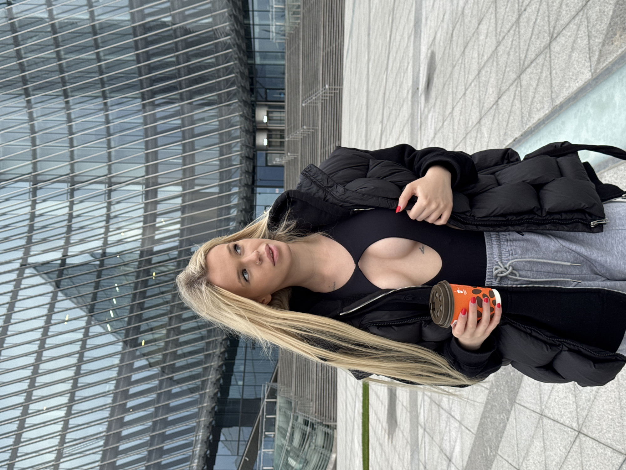 A blonde woman with blue eyes and a nose ring stands outdoors, holding a coffee cup. She is wearing a black puffer jacket over a black top with a cut-out detail, and grey sweatpants. Her nails are painted red. The woman is holding an orange and brown disposable coffee cup with a brown lid. Behind her, a modern building with a glass facade is visible. The scene is set on a paved outdoor area, possibly a plaza or walkway. The lighting suggests it is daytime. The woman's expression appears contemplative or relaxed. There is no visible text in the image. The location is specified as Saint Petersburg, Russia.