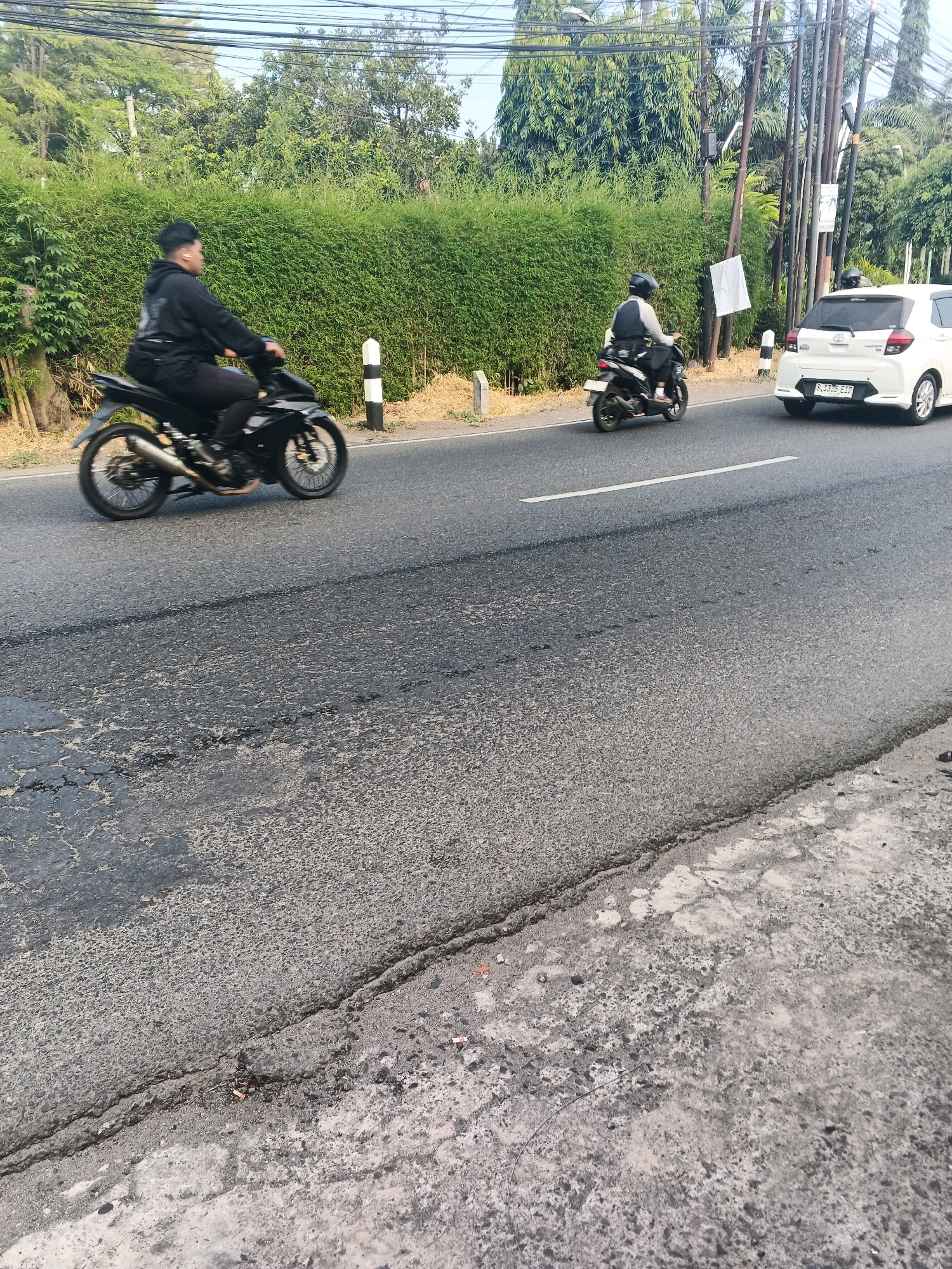In Ungaran, Indonesia, an outdoor scene unfolds on a paved road. Two individuals are riding motorcycles. The person on the left, wearing a black hoodie and pants, is on a black motorcycle. To the right, another person is on a black and white motorcycle, wearing a helmet and a grey and black outfit. A white car is also present on the road. The background features a dense, green hedge row, trees, and utility poles. The road surface shows signs of wear and tear, with visible cracks and uneven patches. The lighting suggests it is daytime, possibly morning or late afternoon, with a clear sky visible.