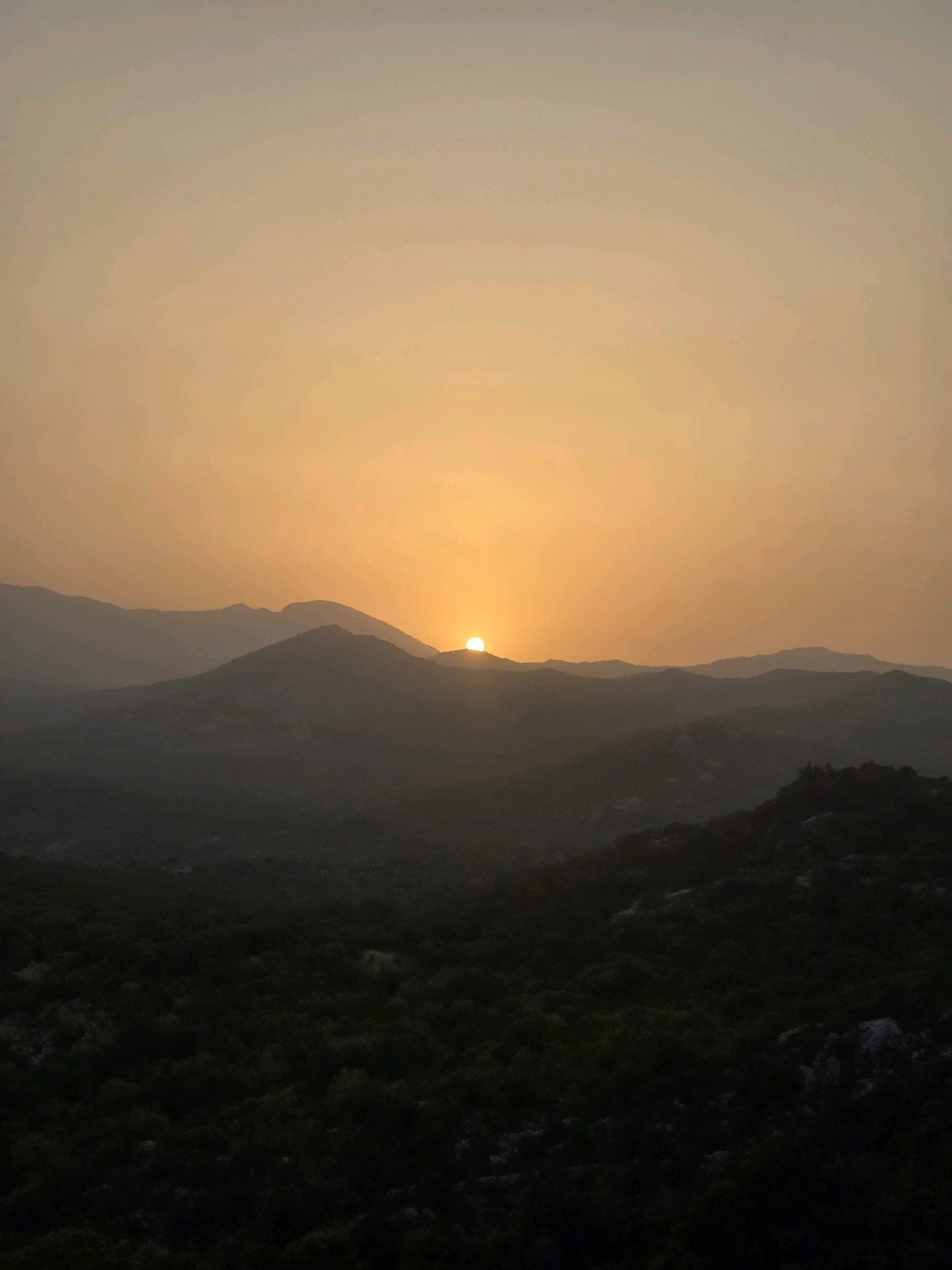 The image depicts a sunset over the mountains surrounding Medun, Montenegro. The sun, a bright orange orb, is partially obscured by the horizon of a distant mountain range. Layers of mountains recede into the background, fading from a darker silhouette to lighter tones due to atmospheric haze. The foreground consists of dark, textured hills covered in dense vegetation, appearing as a forest or scrubland. The sky is a gradient of warm hues, transitioning from a pale yellow near the sun to a muted orange and then a dusty, light brown at the top of the frame. There are no people or man-made structures visible in the image. The overall scene is natural and serene, characterized by the dramatic light of the setting sun casting long shadows and creating a sense of depth.