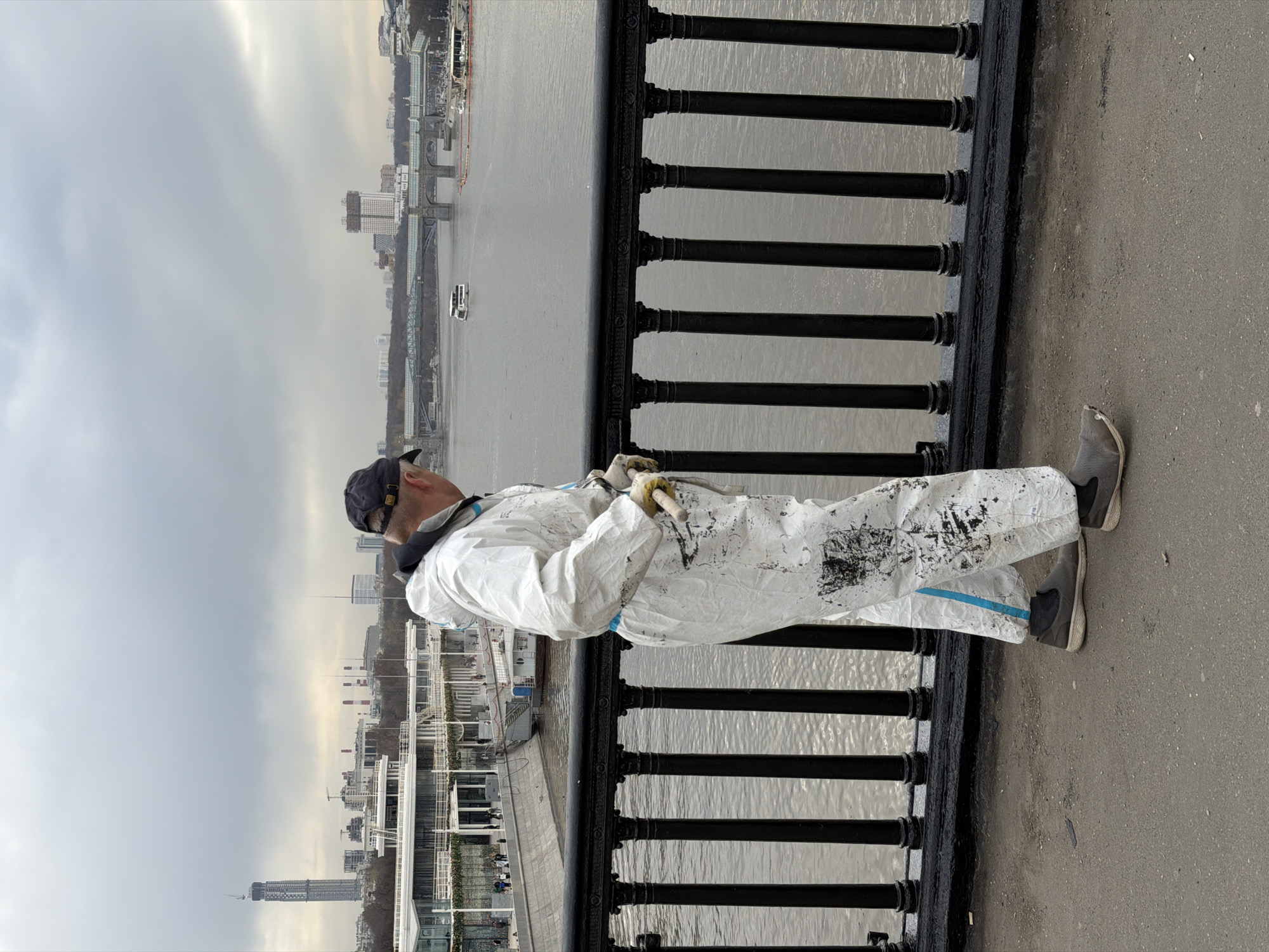 A man in a paint-splattered white jumpsuit and a dark baseball cap is painting a black wrought-iron fence on a bridge in Moscow, Russia. The background shows a wide river and city buildings under a cloudy sky. The man is wearing grey sneakers and yellow gloves. He holds a paintbrush in his right hand.