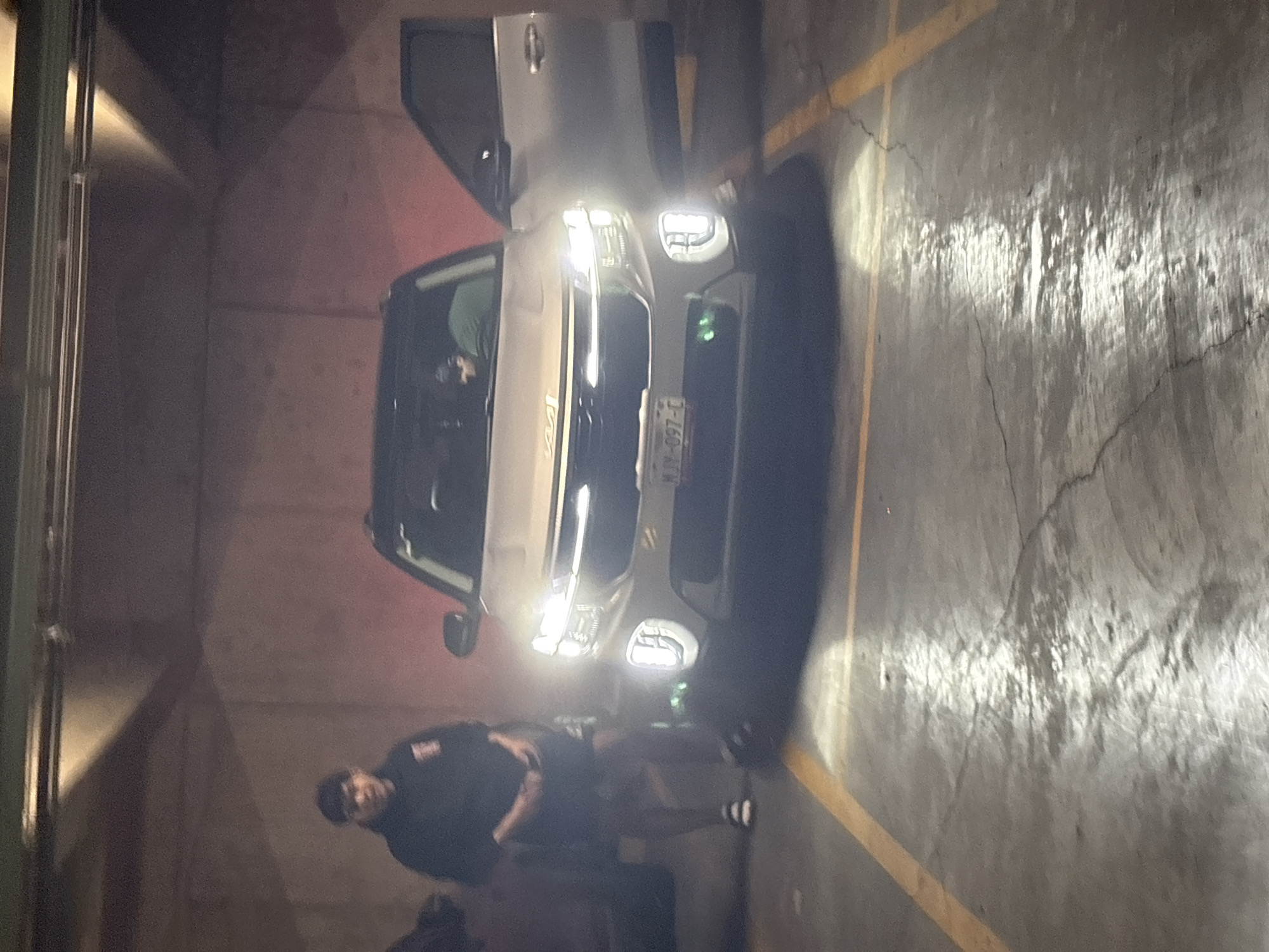 This image depicts a nighttime or dimly lit scene within an indoor parking garage, likely located in Huixquilucan de Degollado, Mexico. The primary subjects are a light-colored car and two individuals.
A white or silver SUV or crossover is prominently featured, with its front headlights and daytime running lights brightly illuminated. The car's driver's side door is open, and a person wearing a green t-shirt is partially visible inside the vehicle, suggesting they are either entering or exiting.
Standing near the front of the car, to the left of the headlights, is another individual. This person is dressed in a dark short-sleeved top and dark pants or shorts, wearing dark shoes with white striped socks. They appear to be leaning forward or looking down, with their posture suggesting a momentary stop or activity.
The setting is a typical concrete parking structure. The walls are made of concrete, possibly with a reddish tint from ambient light or paint, and the floor is concrete with clear yellow parking lines. Cracks are visible on the floor, which is brightly lit by the car's headlamps, creating a strong contrast with the darker surroundings.
A notable detail is the visible license plate on the front of the vehicle, which displays the text "MVN-097-C". The "MVN" prefix is consistent with Mexican license plates, confirming the given location context. The scene captures an ordinary moment of transit or preparation within a parking facility.
