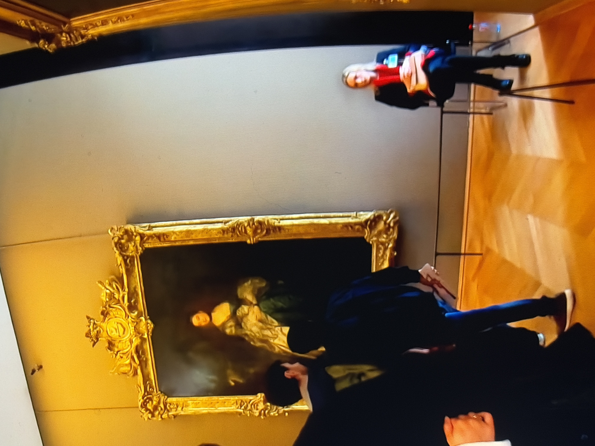 The image depicts an indoor scene, likely a museum or art gallery, with a large, ornate gold-framed painting prominently displayed on a neutral-colored wall. The painting itself features a portrait of a woman in historical attire. In the foreground, three individuals are visible. Two men, seen from behind and the side, stand to the left of the painting, appearing to observe it. The man on the far left is partially obscured. The man in the middle wears a dark jacket and blue jeans, and the man closest to the painting wears a dark hoodie and jeans. To the right of the painting, a blonde woman with a red scarf and a dark jacket is seated on a chair, holding a light-colored bag and looking towards the viewer. A clear plastic bottle is visible on the floor near her. The floor is made of polished wood with a parquet pattern. The overall lighting suggests an indoor environment with artificial light sources, casting warm tones on the walls and floor.