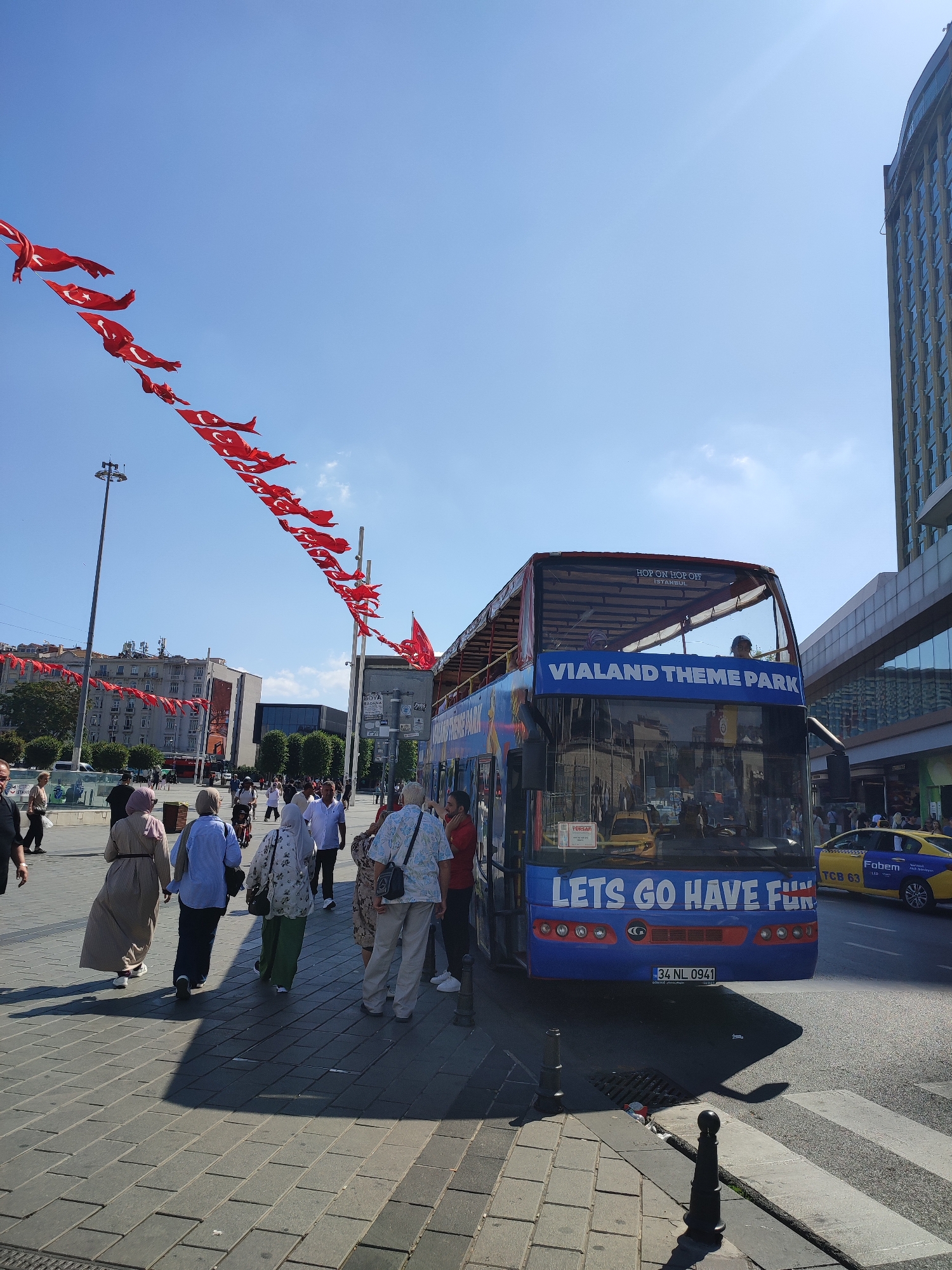 In the city of Beyoğlu, Turkey, on a sunny day, a blue double-decker bus with "Vialand Theme Park" and "Lets Go Have Fun" written on it is parked on a street. Several people are walking on the sidewalk, some are boarding the bus, and others are standing around. Red Turkish flags are strung across the street overhead. A yellow taxi is visible behind the bus. In the background, there are buildings, including a tall modern building. The street is paved with gray cobblestones, and there is a pedestrian crossing.