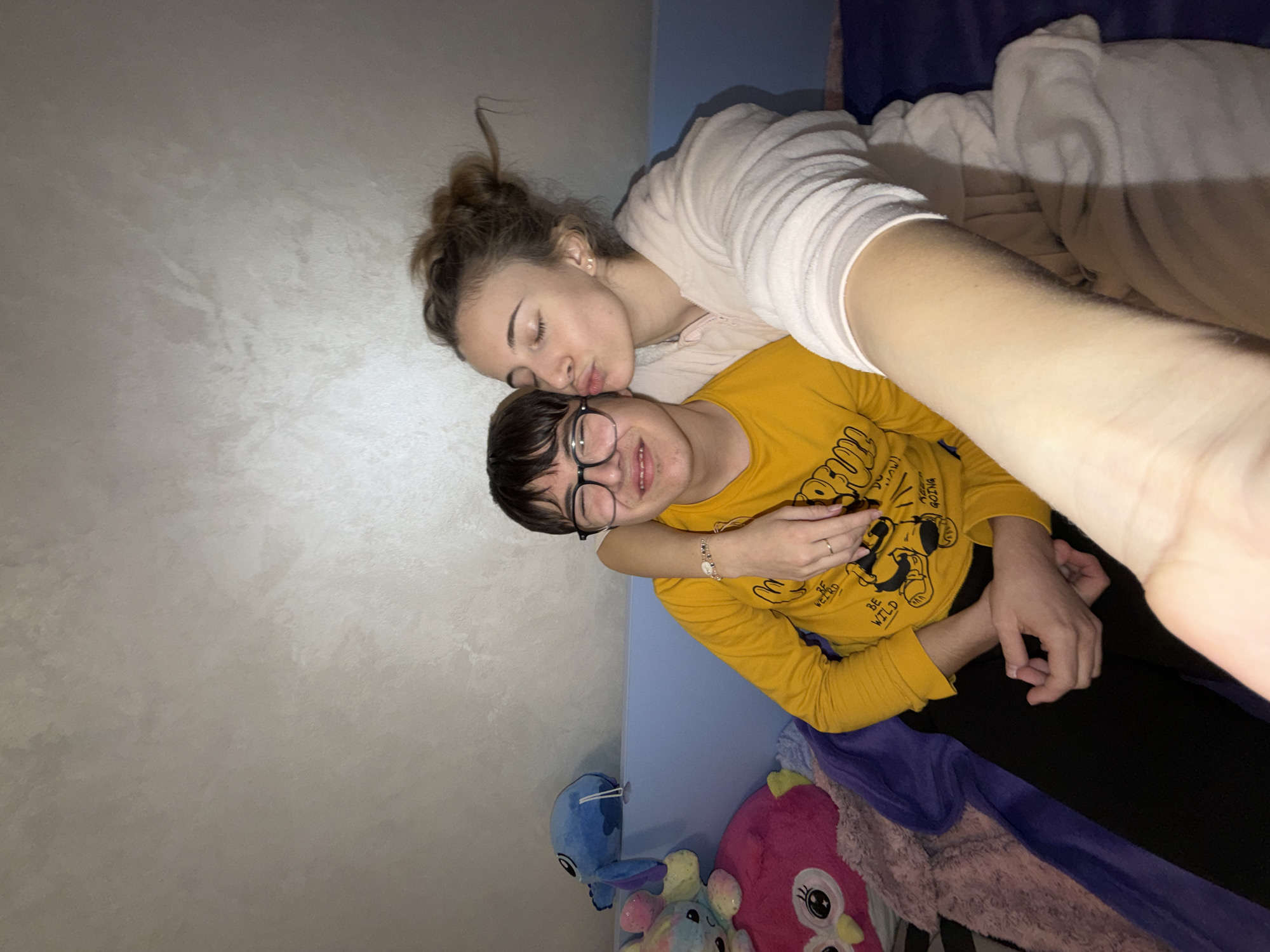 The image depicts two individuals in an indoor setting, likely a bedroom. A male individual, with short dark hair and wearing black-rimmed glasses, a yellow long-sleeved shirt with black graphics, and black pants, is seated on the left. He is smiling with his eyes partially closed. A female individual, with light brown hair styled in a bun, wearing a light pink hooded robe, is positioned to the right and closer to the camera. Her right arm is wrapped around the male's left shoulder, and her hand holds his left hand. Her eyes are closed, and her lips are pursed, making a kissing gesture towards the male's left cheek. The background consists of a light-colored, textured wall and a light blue headboard or bed frame. Plush toys, including a pink owl, a multi-colored creature, and a blue Stitch character, are visible on the bed to the left of the male. Bedding includes a dark blue sheet and a light pink textured blanket. The scene appears to be a selfie taken by the female. The image does not contain identifiable markers to confirm its specific location within Plovdiv, Bulgaria.