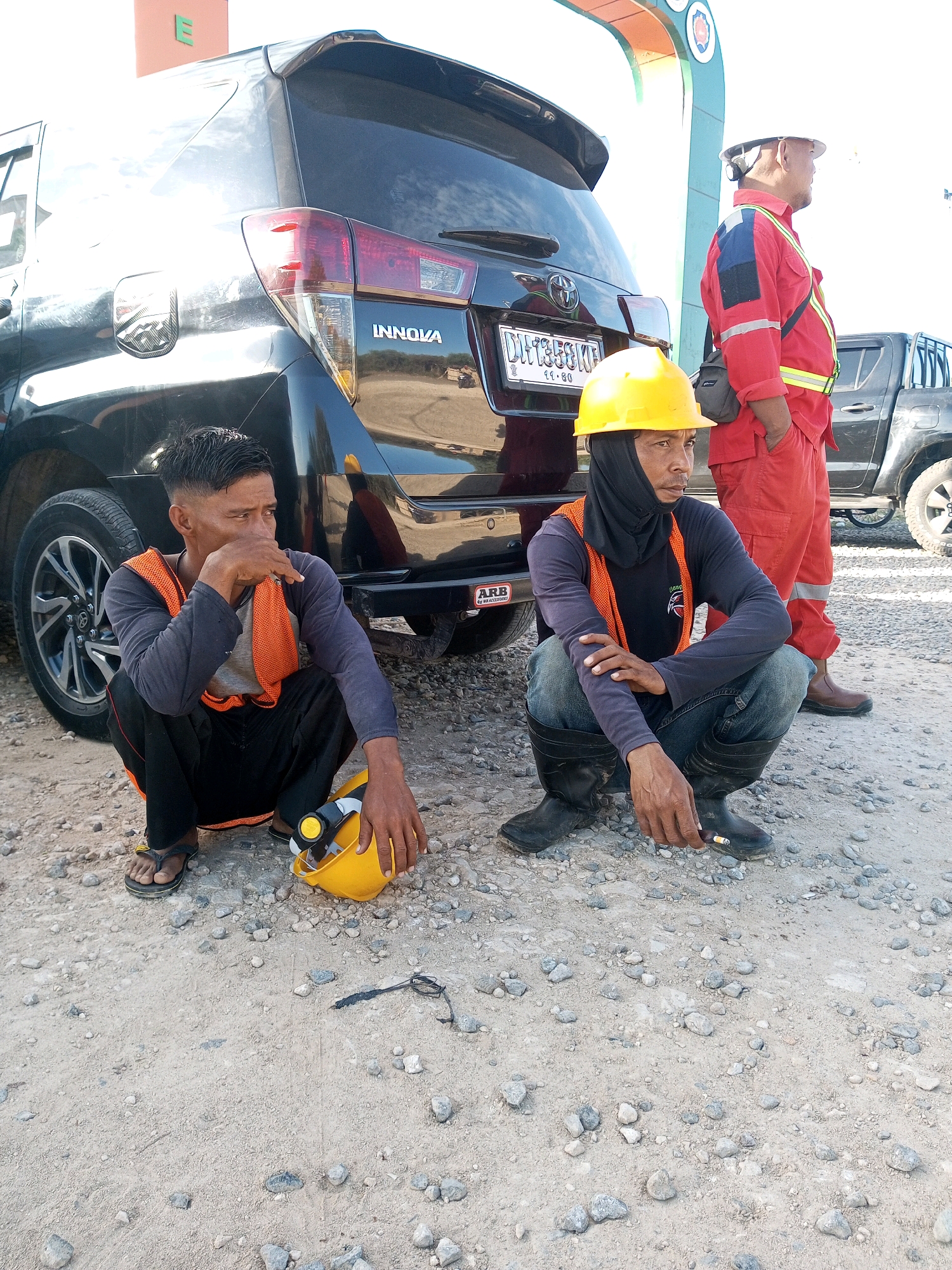 In the city of Matasio, Indonesia, two men are squatting on a gravel surface near a black SUV. The man on the left is wearing a dark long-sleeved shirt, an orange reflective vest, black pants, and flip-flops. He has his hand on his chin, looking to his right. A yellow hard hat is on the ground next to him. The man on the right is wearing a dark long-sleeved shirt, jeans, black rubber boots, and a yellow hard hat. He is holding a cigarette in his right hand and looking to his right. A third man, wearing a red and blue uniform with a reflective yellow stripe and a white hard hat, stands behind the SUV, also looking to his right. The environment appears to be an outdoor industrial or construction area, indicated by the gravel, the vehicles, and the work attire of the individuals. The lighting suggests it is daytime.