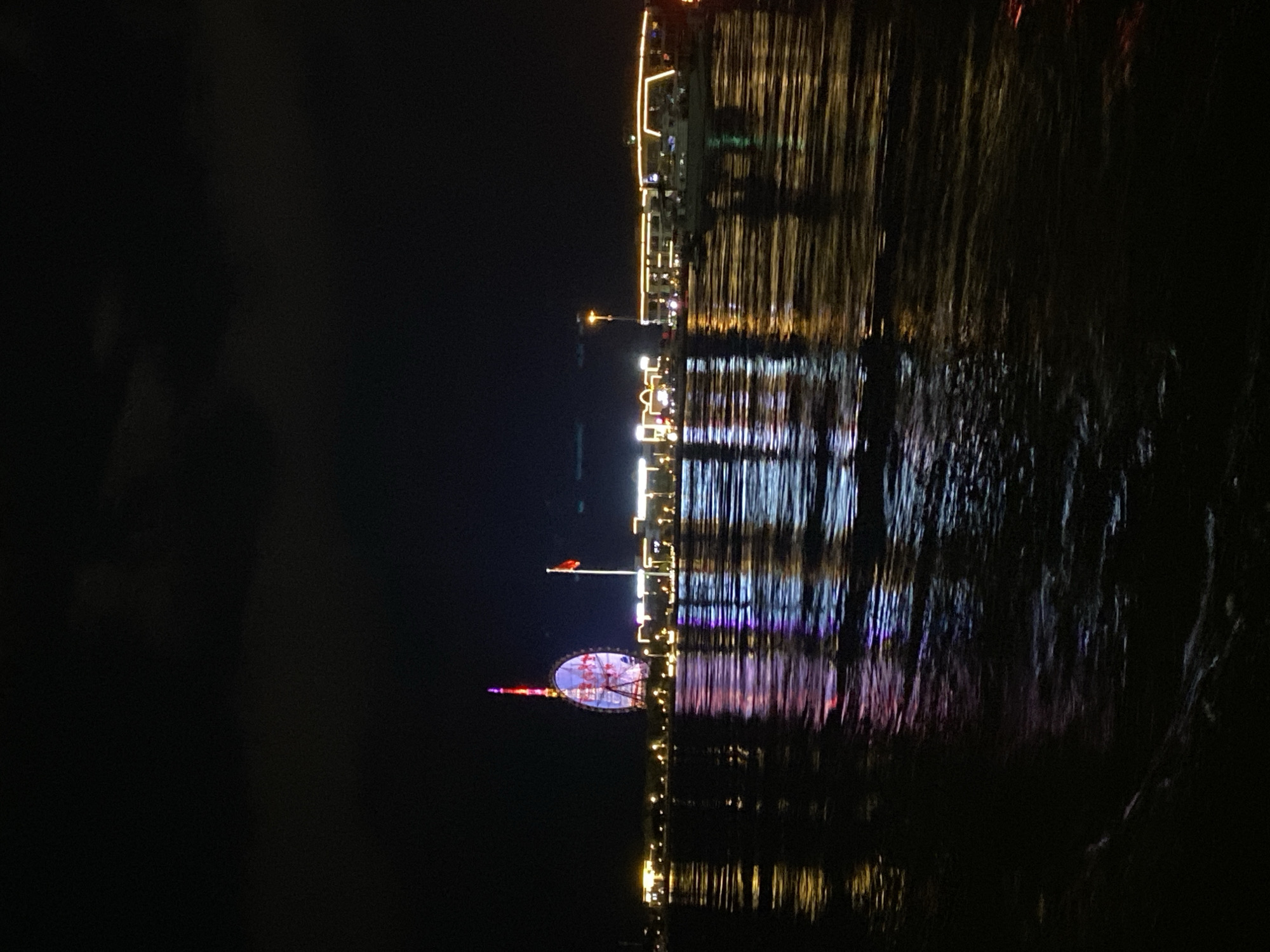 A nighttime photograph captures a wide body of water reflecting the lights of a city skyline. Dominating the left side of the frame is a large, illuminated Ferris wheel, displaying a multi-colored light show with pinks, purples, and blues. To the right of the Ferris wheel, a red flag flies from a pole against the dark sky. Further to the right, a line of buildings is lit with yellow and white lights, and several boats are docked along the waterfront. The water itself is dark, disturbed by ripples that catch and distort the reflections of the city lights, creating streaks of color on its surface. The sky above is mostly black, with some faint clouds visible near the top. The scene appears to be a waterfront area of Blagoveshchensk, Russia, possibly along the Amur River, given the distinct architecture and the presence of the Ferris wheel, a common landmark in such locations.