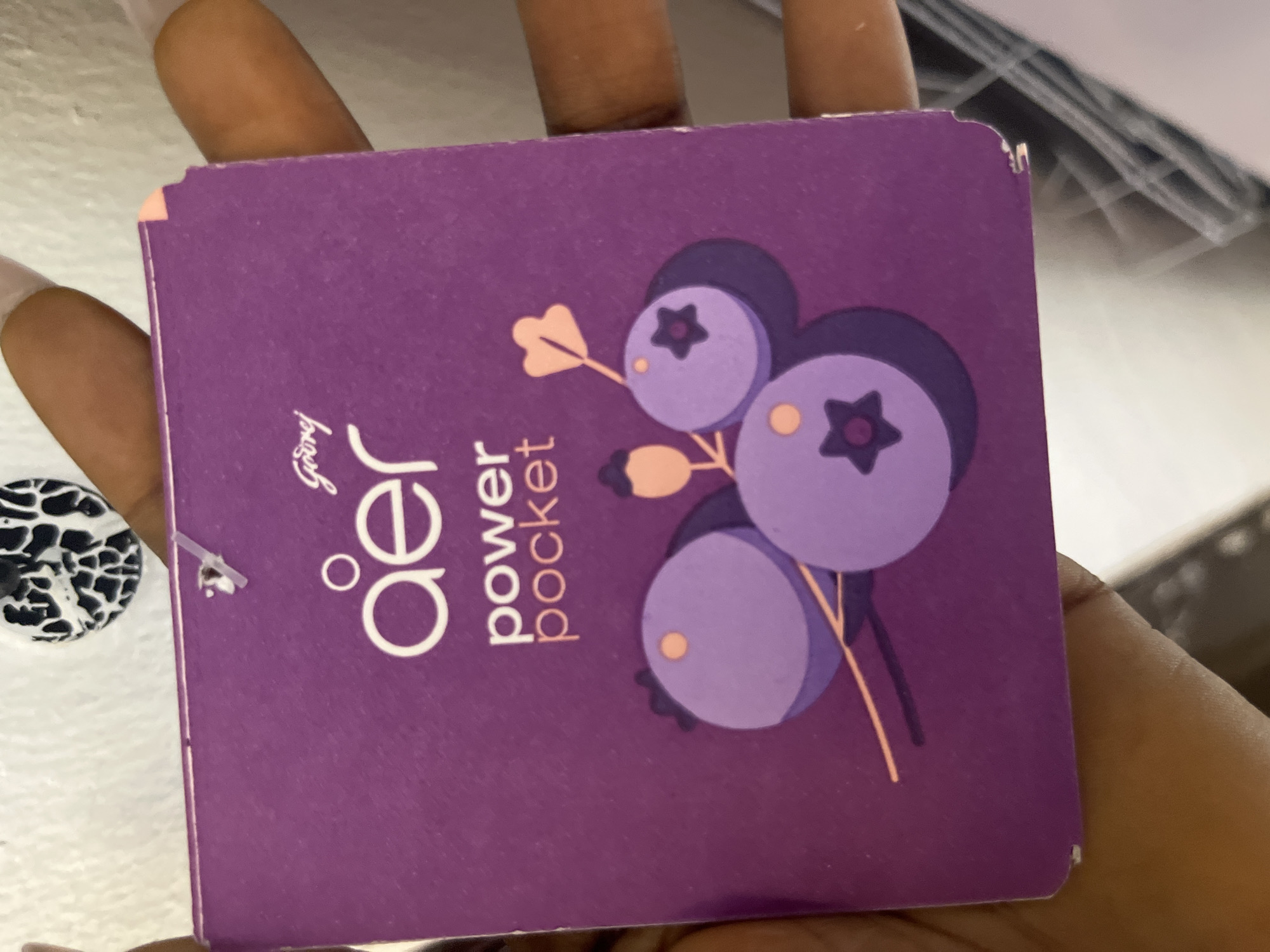A person's hands are holding a square, purple product tag with a white graphic of blueberries. The tag has white text that reads "Godrej" at the top, followed by "aer" and then "power pocket" below. The background is blurred and appears to be a white wall with a black and white patterned object. This image is not from Abuja, Nigeria.