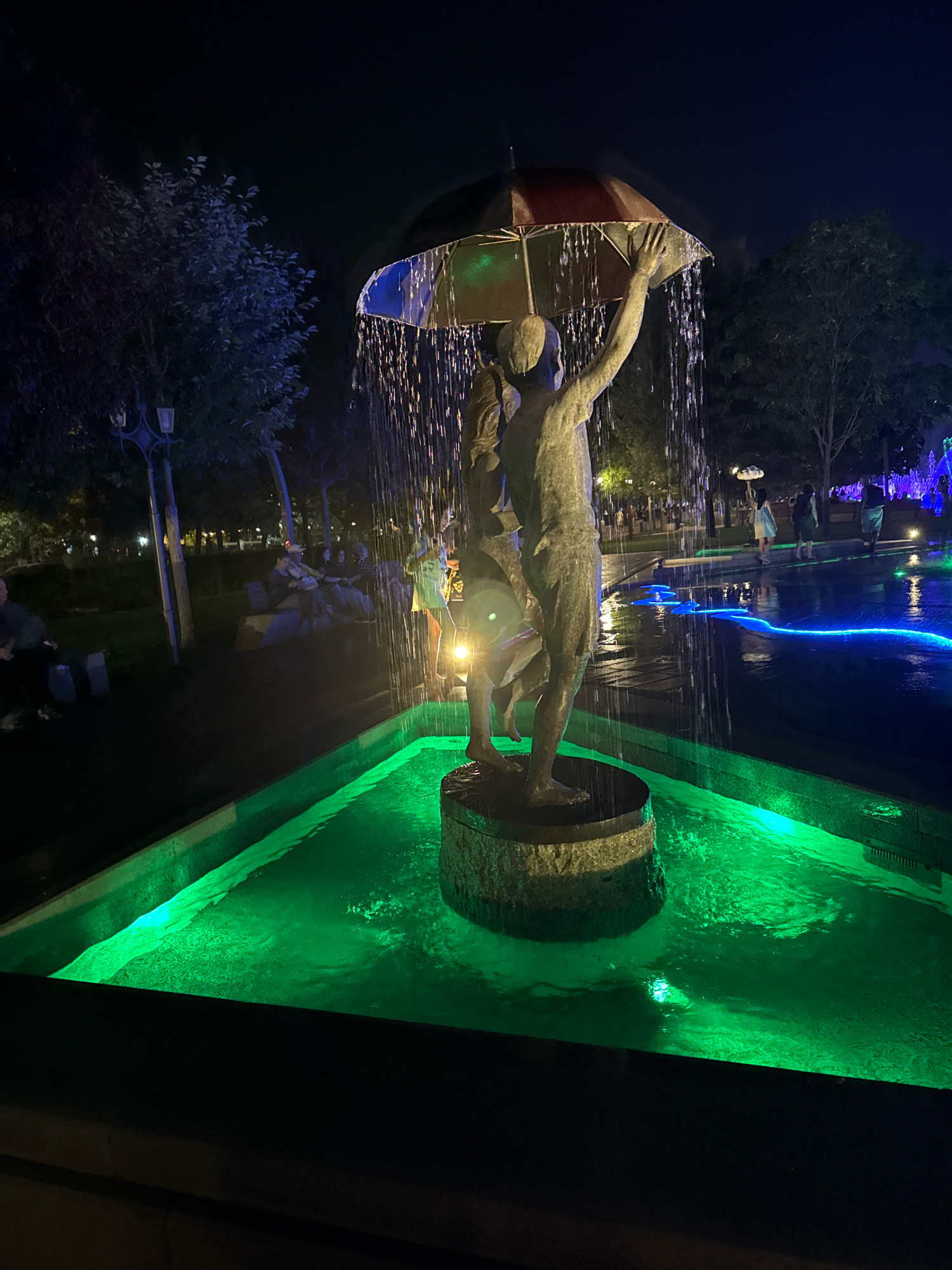 The image shows a nighttime scene in Yerevan, Armenia, featuring a fountain with a sculpture of two figures holding an umbrella. The water from the fountain cascades down, creating a shimmering effect. The fountain pool is illuminated with bright green lights, and the surrounding area is dimly lit with blue lights in a separate water feature in the background. Several trees line the pathway behind the fountain, and a few people are visible sitting on benches and walking in the background, suggesting this is a public park or square. The overall ambiance is atmospheric and somewhat romantic, enhanced by the contrast of the dark night sky with the vibrant illumination of the fountain.