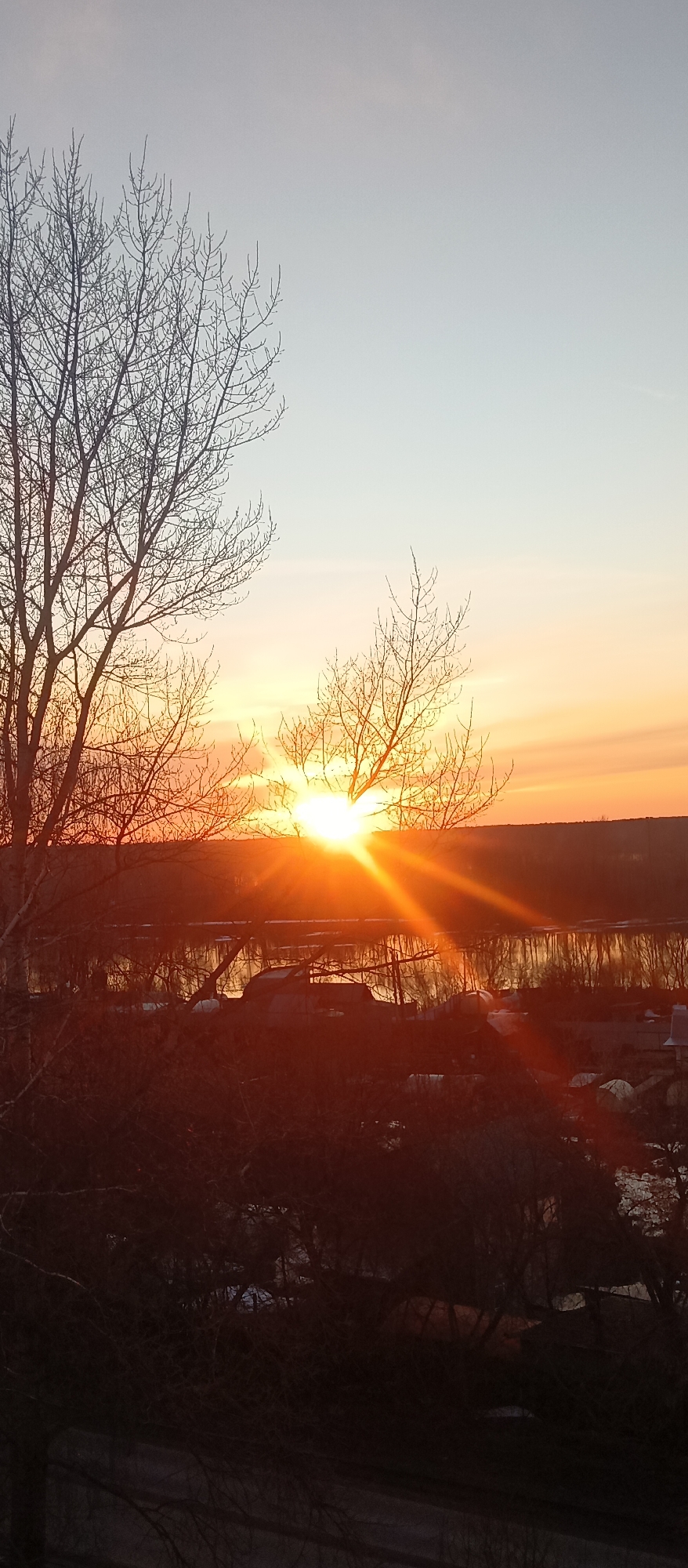 The image shows a sunset over a body of water, likely a river or lake, in an urban or suburban environment. The sun is partially obscured by bare trees, creating a burst of light and visible rays. The sky transitions from a pale blue to shades of orange and yellow near the horizon. The body of water reflects the colors of the sunset. The foreground is filled with dark, silhouetted trees and vegetation, and some indistinct structures that appear to be buildings. The location is Unknown, Unknown.