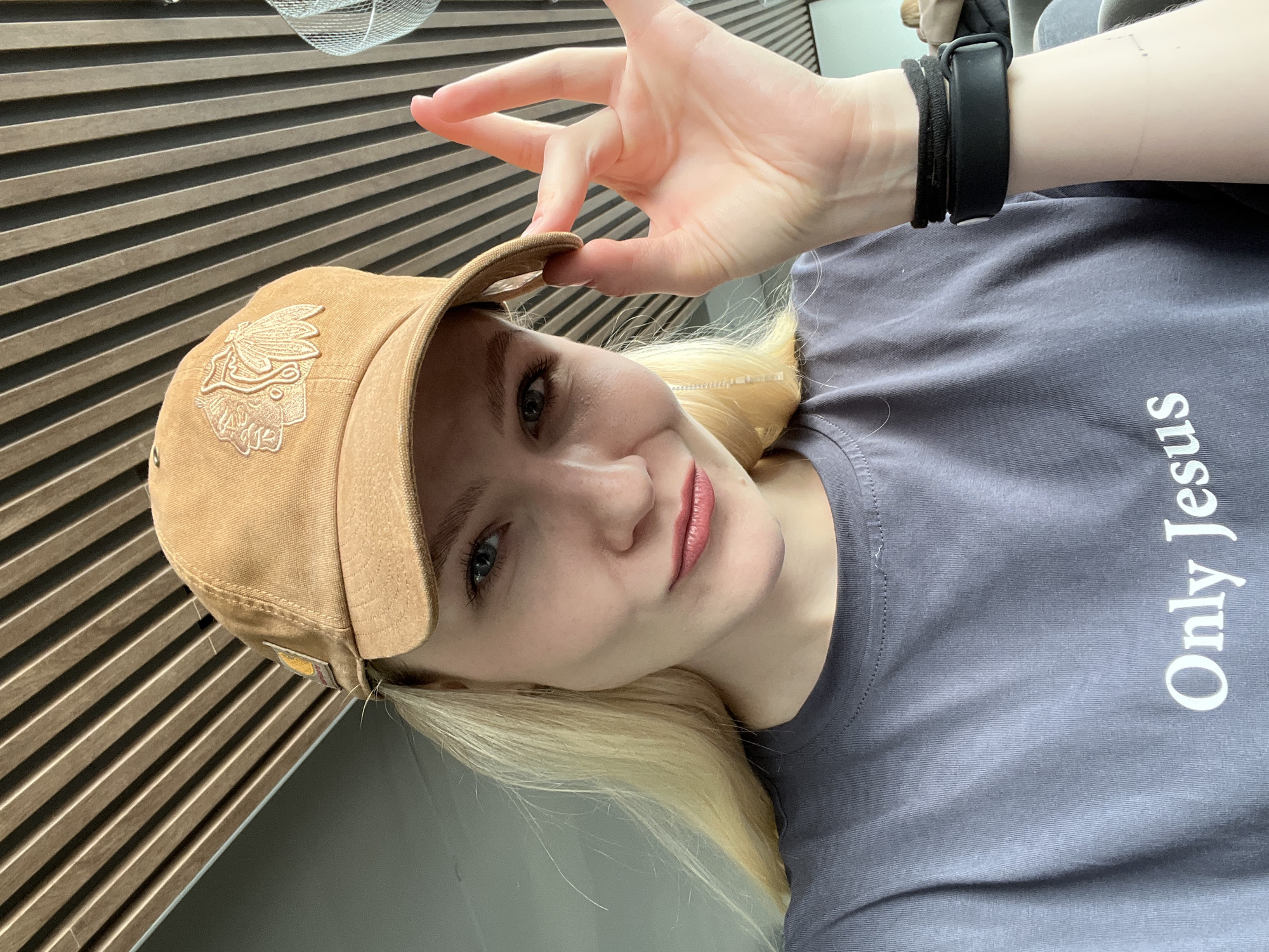A person with blonde hair is wearing a light brown baseball cap with an embroidered design on the front and a gray T-shirt with the words "Only Jesus" printed vertically in white on the right side of the shirt. A black fitness tracker is on the person's wrist, and the person is using their left hand to adjust the cap. The background consists of a wall with horizontal brown slats. The photo was likely taken indoors. The location information, Khimki, Russia, is not apparent from the image.