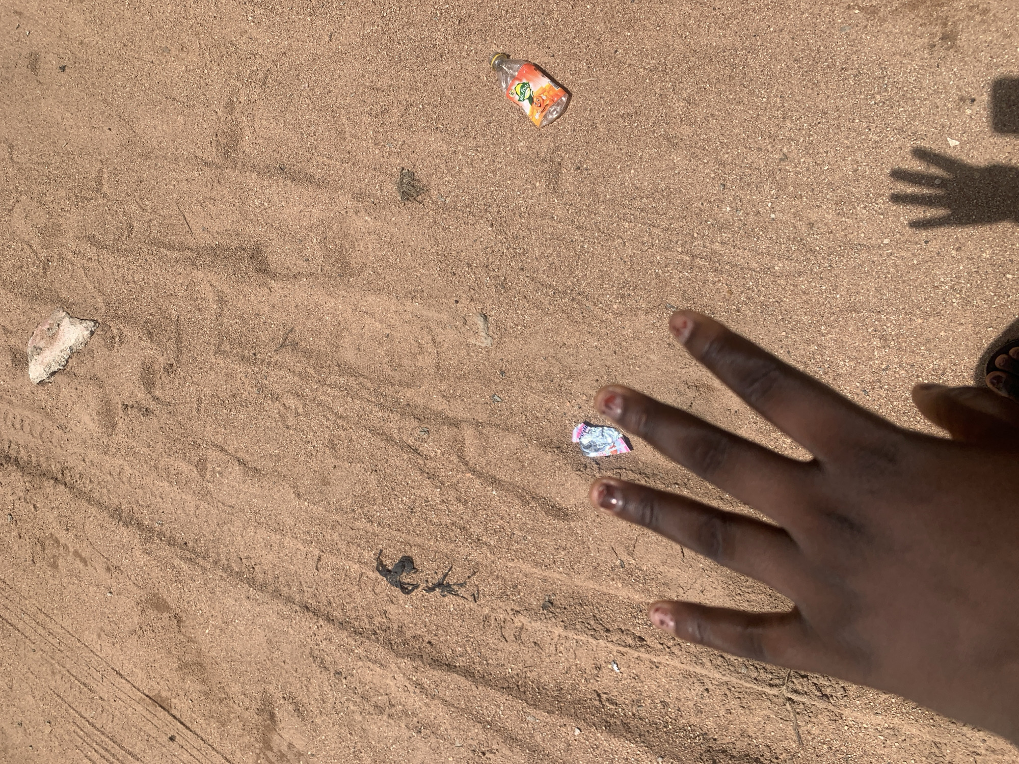 A close-up, overhead shot captures a person's dark-skinned hand with painted fingernails reaching into the frame, obscuring a portion of the sandy ground in Jauro Yinu, Nigeria. The ground is covered in fine, light brown sand, with faint tire tracks visible on the left side of the image. Several pieces of litter are scattered across the sand: a small, crumpled wrapper near the fingers, a piece of light-colored rock in the upper right quadrant, and a crushed plastic bottle with an orange and yellow label that reads "Mr. Fruit" to the right of center. A dark, organic clump, possibly dried vegetation or animal remains, is also visible in the upper left quadrant. The lighting suggests a sunny day, casting a shadow of the hand and a partial shadow of a shoe on the ground.