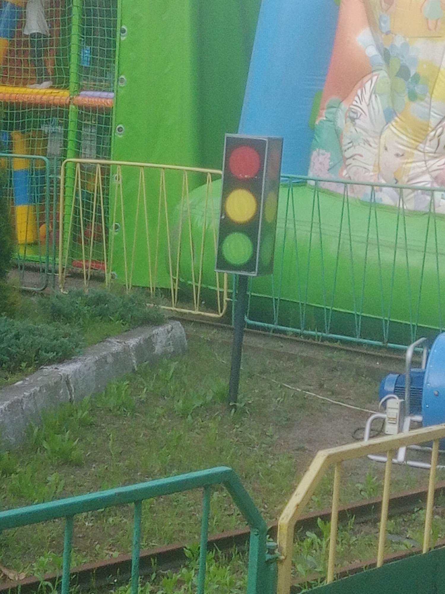 A traffic signal with red, yellow, and green lights is positioned in front of a green inflatable structure. The traffic signal is tall and black with circular lights. The red light is at the top, followed by yellow, and then green. The traffic signal is mounted on a pole. Behind the traffic signal, there is a large green inflatable structure. To the left of the traffic signal, there is a yellow metal fence and a netted structure with colorful bars. In the foreground, there is a concrete barrier and green grass. To the right of the traffic signal, there is a blue piece of machinery. This scene is located at an amusement park in Horishni Plavni, Ukraine.