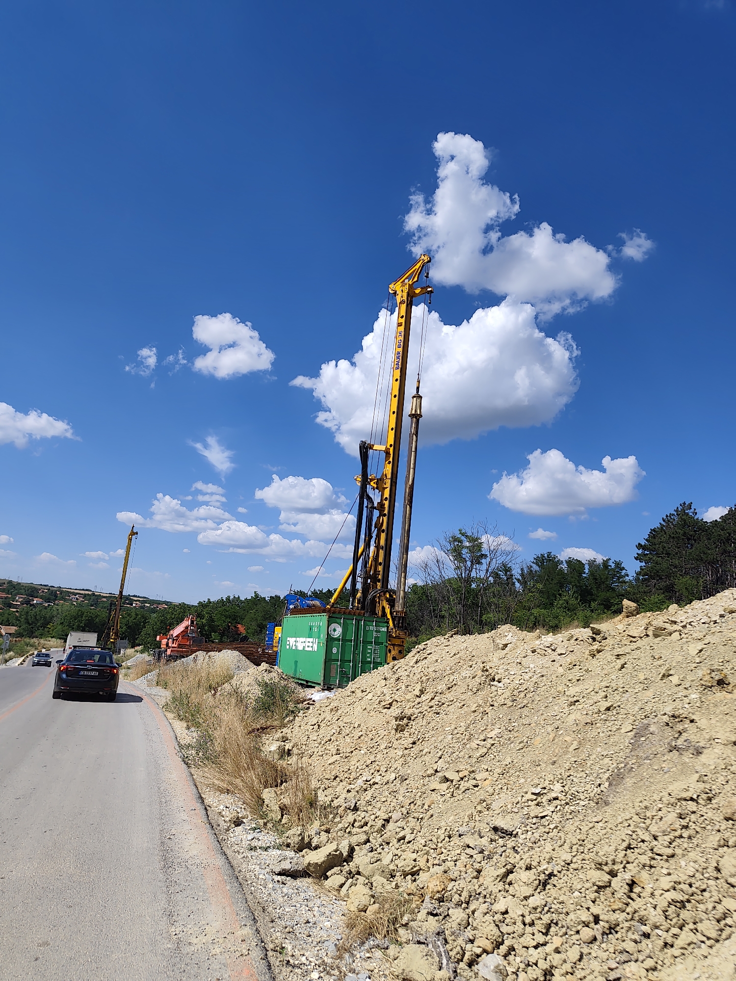 This image captures a construction scene on a sunny day in Sadovets, Bulgaria. A large yellow drilling rig, identified as a BAUER BG 36, is positioned prominently on the right side of the frame, next to a pile of dirt and rocks. To the left, a road curves into the distance, with two cars driving on it. In the background, a line of trees and some houses suggest a semi-urban or rural setting. The sky is a vibrant blue, dotted with fluffy white clouds. A green shipping container is visible near the base of the drilling rig, labeled "EVERGREEN". The overall impression is one of ongoing infrastructure development in a Bulgarian town.