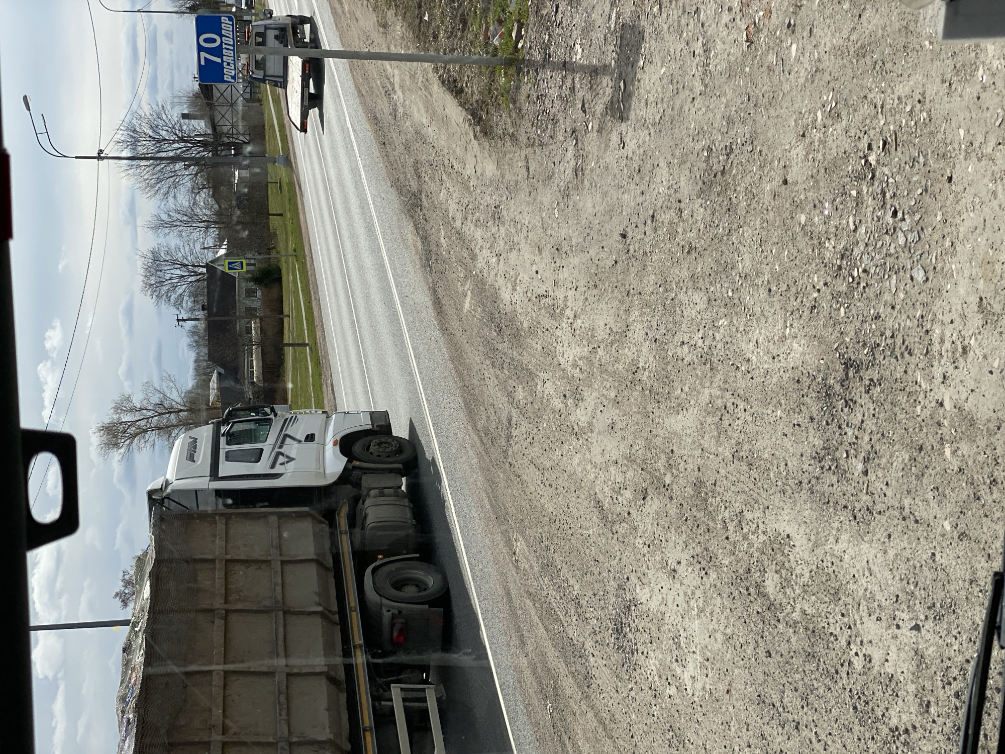 The image shows a road in Bol'shoe Teshkovo, Russia. A large white truck carrying a load of concrete blocks is driving on the road. A speed limit sign indicating "70" in Russian is visible along the road. The surrounding environment consists of trees, buildings, and a dirt shoulder next to the road. The image appears to be taken from inside a vehicle, possibly another truck.
