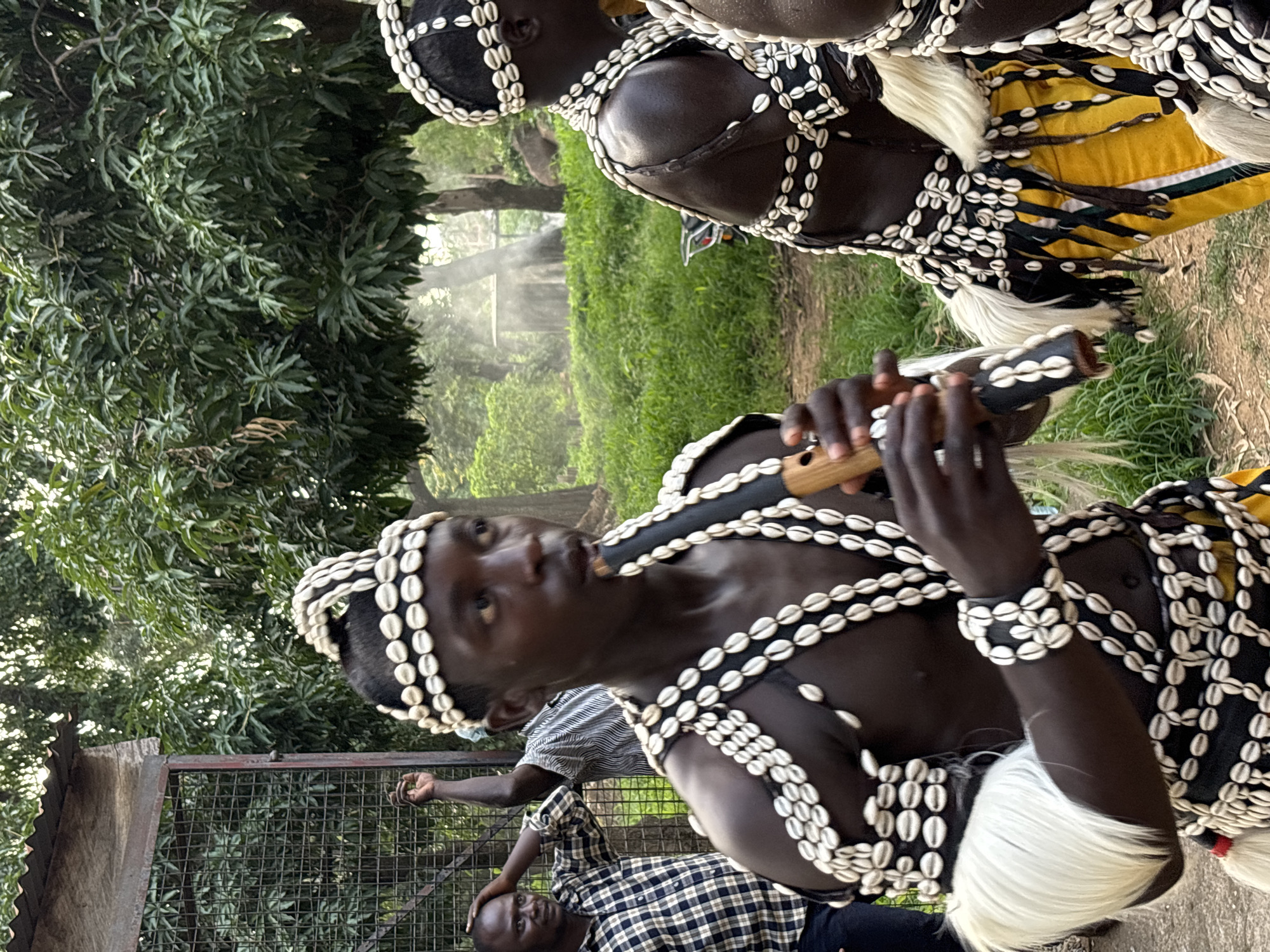 This is a medium shot photograph of a group of people in an outdoor setting, possibly a village or a cultural event. In the foreground, a young Black man, likely a performer, is playing a wooden flute. He is adorned in a traditional costume consisting of a headdress, necklaces, bracelets, and armbands made of numerous white cowrie shells. His chest and arms are bare, and he wears a skirt-like garment with yellow and black fabric and white fur accents. Behind him and to the left, another Black man in a checkered shirt is seen with his hand on his head, looking towards the performer. Further back, another man in a striped shirt stands with his arm raised. The background features lush green vegetation, trees with dense foliage, and a rustic structure that appears to be a fence or enclosure. Sunlight filters through the trees, creating a soft, diffused light with some visible haze or mist. The ground is covered with dirt and possibly fallen leaves. The scene has a warm, natural ambiance, suggesting it is daytime. The location is identified as Jos, Nigeria.