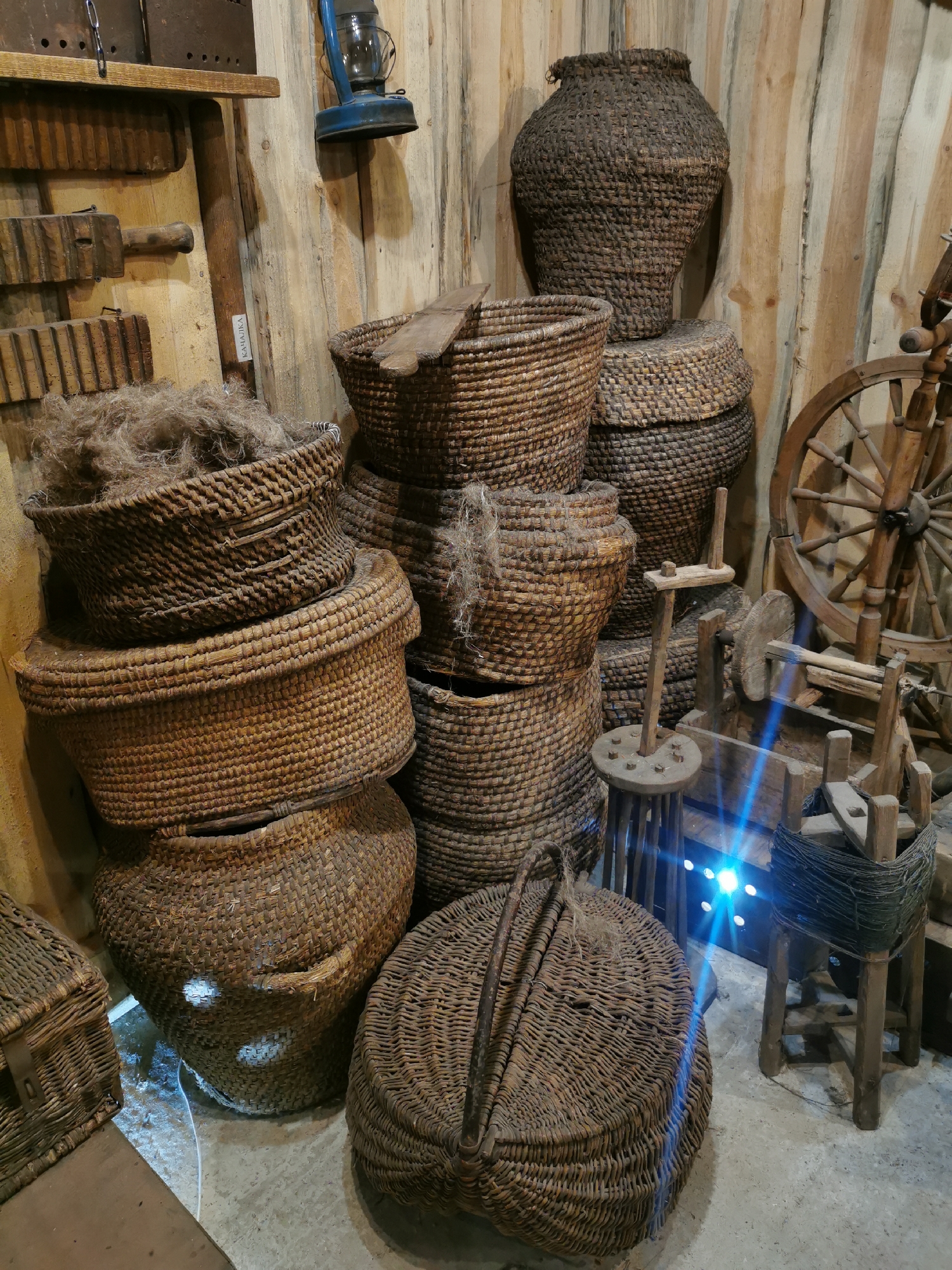 The image depicts a rustic collection of woven baskets and antique tools displayed against a wooden plank wall. In the foreground, a large, round, lidded basket with two handles sits on the concrete floor. To its left, a smaller, rectangular woven basket is partially visible. A stack of woven baskets of varying sizes is arranged in the center and right of the image. The largest basket in this stack is a tall, dark, amphora-shaped piece. To the right, a wooden spinning wheel and other unidentified wooden contraptions are positioned. A blue kerosene lantern hangs on the wall above a wooden shelf, on which several wooden implements are resting. The overall scene suggests a historical or ethnographic display, likely in a rural setting. The presence of a label with the text "КАЛІНКА" on a wooden board hints at a possible Belarusian origin, further supported by the mention of Шчарчоўскі сельскі Савет, Belarus, which is a rural administrative unit. The lighting in the scene is moderately dim, with some artificial light sources casting blue hues on the floor and some of the objects. There are no people visible in the image.