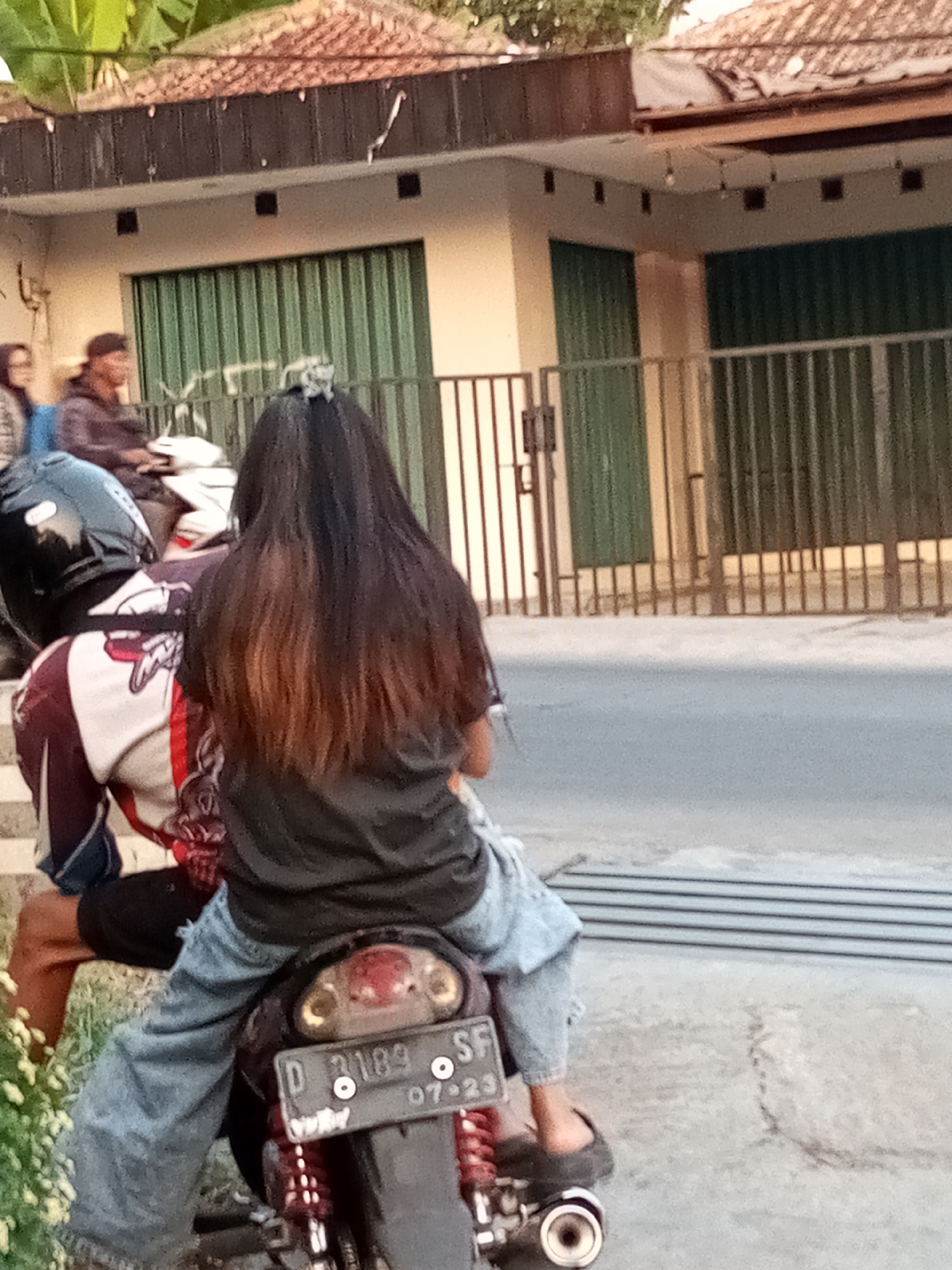 The image shows two individuals on a motorcycle parked on the side of a street in Sariwangi, Indonesia. The motorcycle has a license plate that reads "D 3189 SF 07-23". The person in the front, wearing a black helmet, has a jacket with red and white patterns and appears to be male. The person in the back has long, dark hair with reddish-brown highlights and is wearing a black t-shirt and blue jeans. They are seated behind the driver. The background features a building with green roll-up doors and a tiled roof, typical of Indonesian architecture. A metal fence runs along the front of the building. There are also some plants and a paved surface in the foreground and on the street. The lighting suggests it is daytime, possibly late afternoon given the shadows.