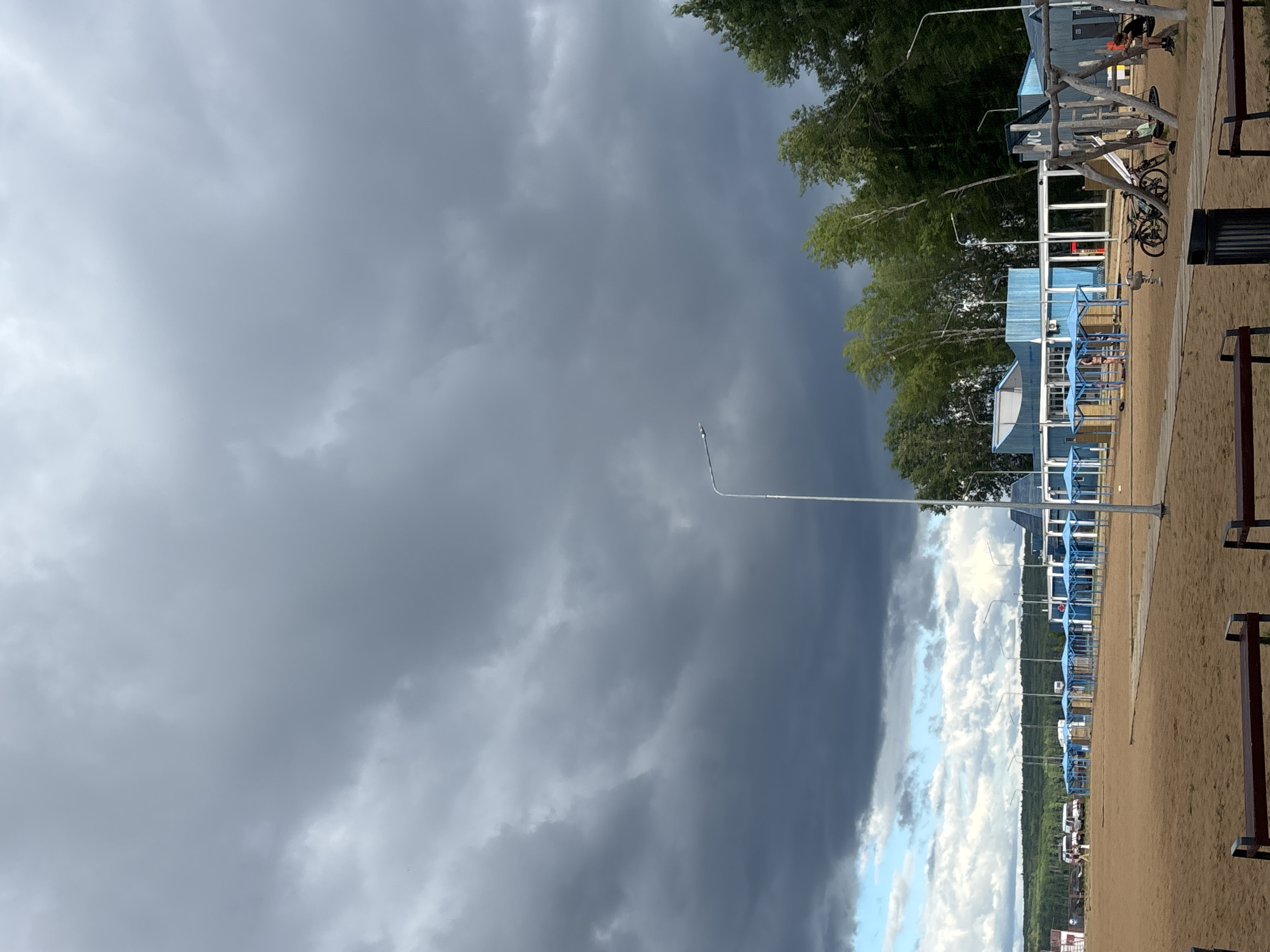 This media file depicts a beach scene in Chaikovsky, Russia. The image is oriented vertically, with the sky dominating the left side of the frame and the beach and structures on the right. Dark, stormy clouds cover a large portion of the sky, suggesting an impending storm or recent rainfall. However, patches of lighter clouds and a sliver of blue sky can be seen towards the right, indicating some variation in the weather.
On the right side of the image, a sandy beach stretches into the distance. Several blue and white structures with open fronts, resembling beach pavilions or changing rooms, are lined up along the shore. Trees with lush green foliage are visible behind these structures, forming a treeline that separates the beach area from what appears to be a more distant landscape or water body.
There are a few people visible in the scene. One person is seated on a bench near some outdoor fitness equipment, and another is engaged in an activity near a bicycle. Two bicycles are parked near one of the structures. The presence of benches and fitness equipment suggests this is a recreational area. The overall mood of the image is somewhat dramatic due to the stormy sky, contrasted with the orderly arrangement of the beach facilities. The time of day is unclear due to the overcast conditions. There is no visible text in the media file.