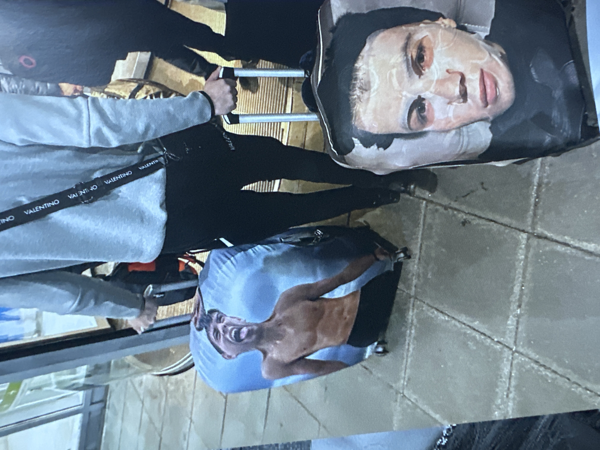 In Sofia, Bulgaria, two individuals are seen on a paved outdoor area, likely near a building entrance with glass doors. One person, wearing a grey hoodie and black pants, is holding the handle of a wheeled suitcase. This suitcase is covered with a print of a shirtless young man with an open mouth and outstretched tongue against a sky-blue background. Next to them, another suitcase features a close-up print of a person's face wearing a white sheet mask. The overall scene depicts individuals with their luggage in an urban outdoor setting.