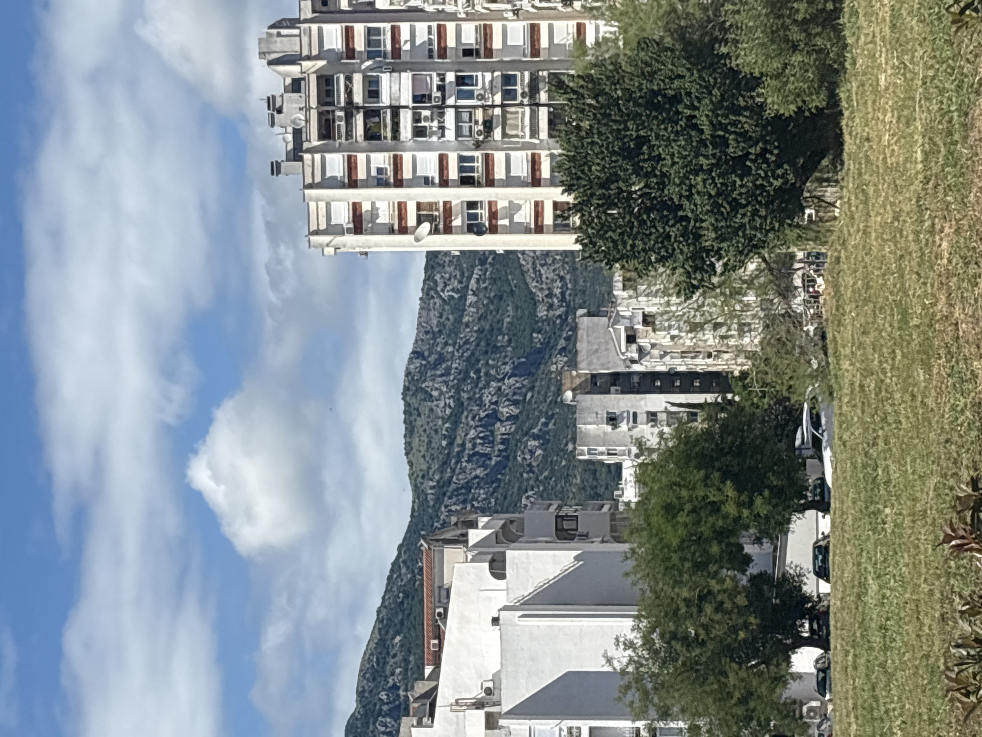 The image captures a vibrant urban scene nestled against a mountainous backdrop, likely in Bar, Montenegro, given the context. The photograph is taken during daytime under a bright blue sky dotted with scattered white clouds.
In the foreground, a strip of green grass stretches across the bottom, with some lush green trees and bushes partially obscuring the view. A street is visible between the greenery and the buildings, with a few cars parked or driving, though not in sharp focus.
The midground is dominated by several multi-story buildings. A prominent, tall apartment building with a white facade and dark red/brown accents stands out. It features numerous windows, balconies, external air conditioning units, and satellite dishes, suggesting a residential or mixed-use purpose. Other white buildings, some with stepped or terraced designs, are visible further back and to the left, also equipped with balconies and AC units. Green trees are interspersed among these structures, adding natural elements to the urban environment.
In the background, a large, rugged mountain range rises dramatically, its slopes covered in dark green and grey foliage and rock formations, creating a striking natural boundary for the settlement.
No people are clearly visible, and no specific activities are taking place. The overall impression is one of a modern town situated in a scenic coastal or mountainous region. There is no visible text in the image.