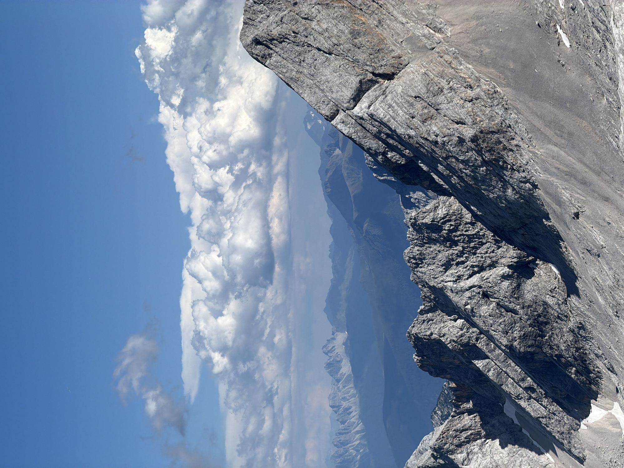 The image depicts a panoramic view of rugged, mountainous terrain under a bright blue sky dotted with fluffy white clouds. The foreground is dominated by the steep, rocky slopes of a mountain, characterized by sharp, jagged peaks and deep shadows. The rock faces exhibit textured layers and are interspersed with patches of snow in the crevices.
In the mid-ground, a range of further mountains recedes into the distance, appearing progressively softer and bluer due to atmospheric perspective. These mountains are also characterized by their pointed peaks and sloping sides, creating a layered effect.
The sky above is a vibrant blue, with large, cumulus clouds forming a dramatic backdrop. The sunlight casts distinct shadows on the mountains, highlighting their dramatic contours. There are no visible people, animals, or man-made structures in the scene. The environment is entirely natural, showcasing the raw beauty of high-altitude alpine landscapes.
Given the context provided, this scene is likely located in the vicinity of Garmisch-Partenkirchen, Germany, a region renowned for its dramatic mountain scenery, particularly the Wetterstein Alps. The view is consistent with vantage points offering expansive vistas of these iconic German peaks.
