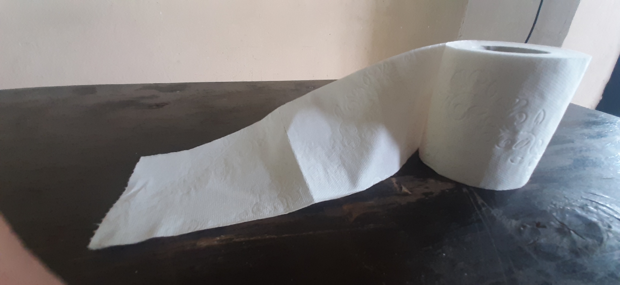 A roll of toilet paper is depicted on a dark, wooden table, with its paper unspooled and trailing across the surface. The toilet paper is white and has a textured pattern. The table appears somewhat worn and possibly covered with a protective sheet. In the background, a wall painted in a light, neutral color is visible. The lighting suggests it could be daytime, with natural light sources illuminating the scene. There are no people visible in the image, and the setting is indoors, likely a room in a home or similar environment. The focus is entirely on the toilet paper roll and its unspooled paper. There is no visible text in the image.