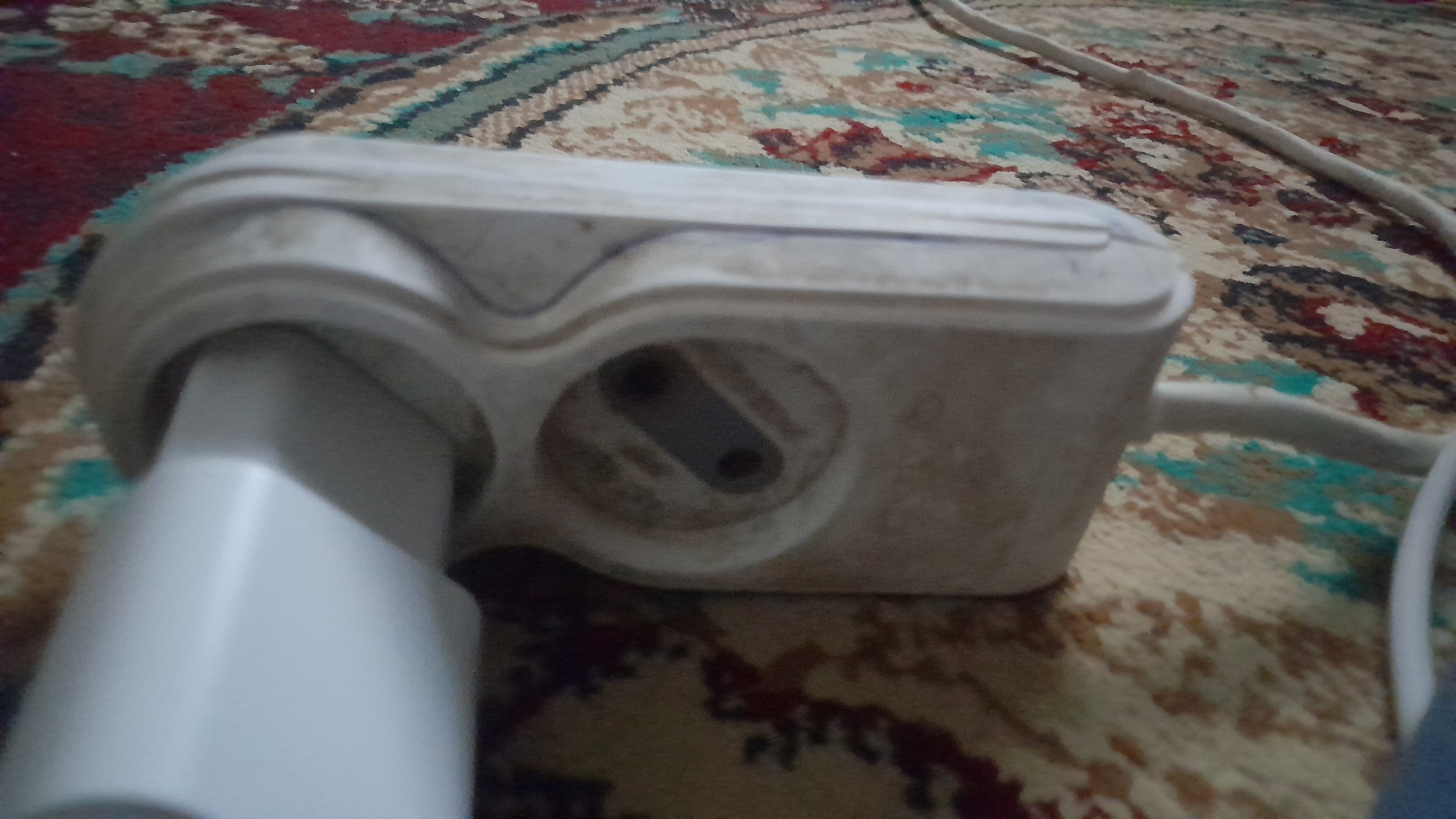 A white, multi-socket electrical adapter is plugged into an electrical outlet. The adapter is a light beige color and has two sockets. One socket appears to be a standard type, while the other is a flatter, wider design. A white electrical plug is inserted into the standard socket. The adapter and plug are resting on a patterned carpet with red, brown, and teal designs. A white electrical cord extends from the adapter and runs across the carpet. The scene is indoors, likely in a residential setting. No individuals are visible in the image. The location is not specified in the image.