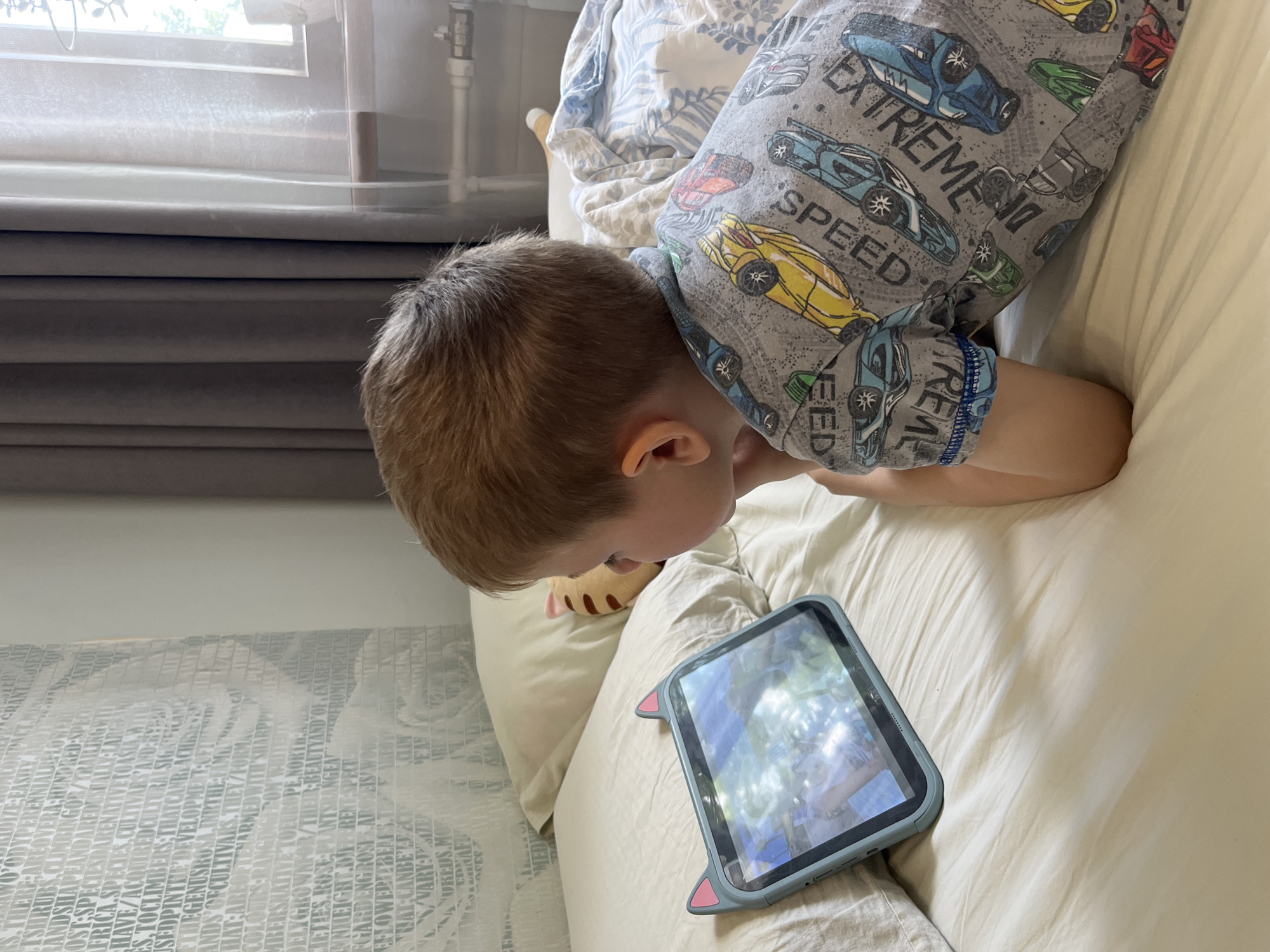 A young child lies on a bed, facing a tablet. The child has light brown hair and is wearing a grey t-shirt with a pattern of cars and text that reads "SPEED" and "EXTREME". The child's arms are resting on the bed. The tablet is in a light blue case with cat ears on top. The screen of the tablet shows a colorful image. The bed is covered with a white duvet. Behind the child, there are pillows and a wall with a patterned wallpaper. The room is lit by natural light coming from a window, partially covered by curtains. The image is taken from a slightly elevated angle.