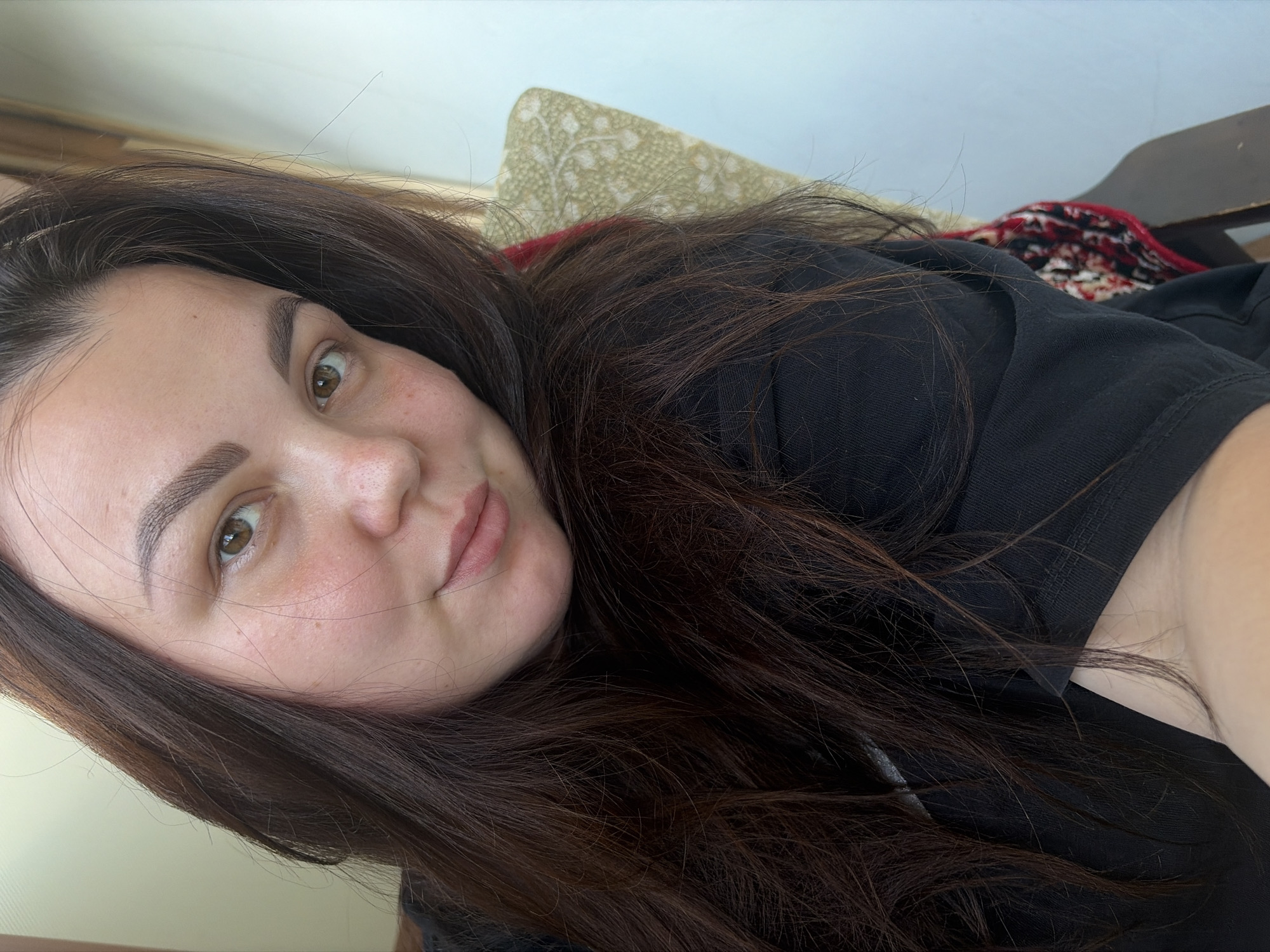 The image is a close-up selfie of a young woman with dark brown hair and light brown eyes. She is wearing a black t-shirt and is seated in a chair with a patterned cushion behind her. The lighting suggests an indoor environment, possibly near a window. There is no indication of the specific location in Izhevsk, Russia.