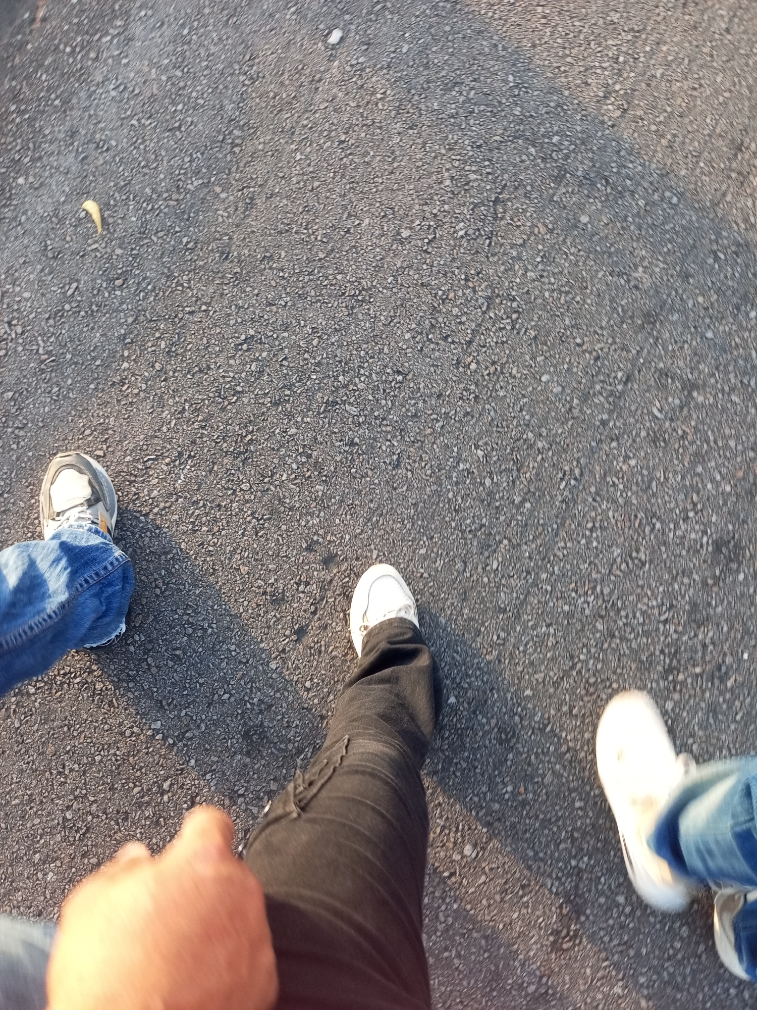 The image depicts a section of asphalt pavement, possibly a road or parking lot, and the lower portions of three individuals. The person on the left is wearing blue jeans and white and gray sneakers. The individual in the center is wearing black pants and white sneakers. The person on the right wears blue jeans and white sneakers. The picture appears to be taken at ground level, suggesting a first-person perspective while walking. A small, yellow crescent, possibly a leaf or trash, is visible on the pavement in the upper left portion of the frame. No other indicators suggest a specific location, such as Juan Griego, Venezuela.
