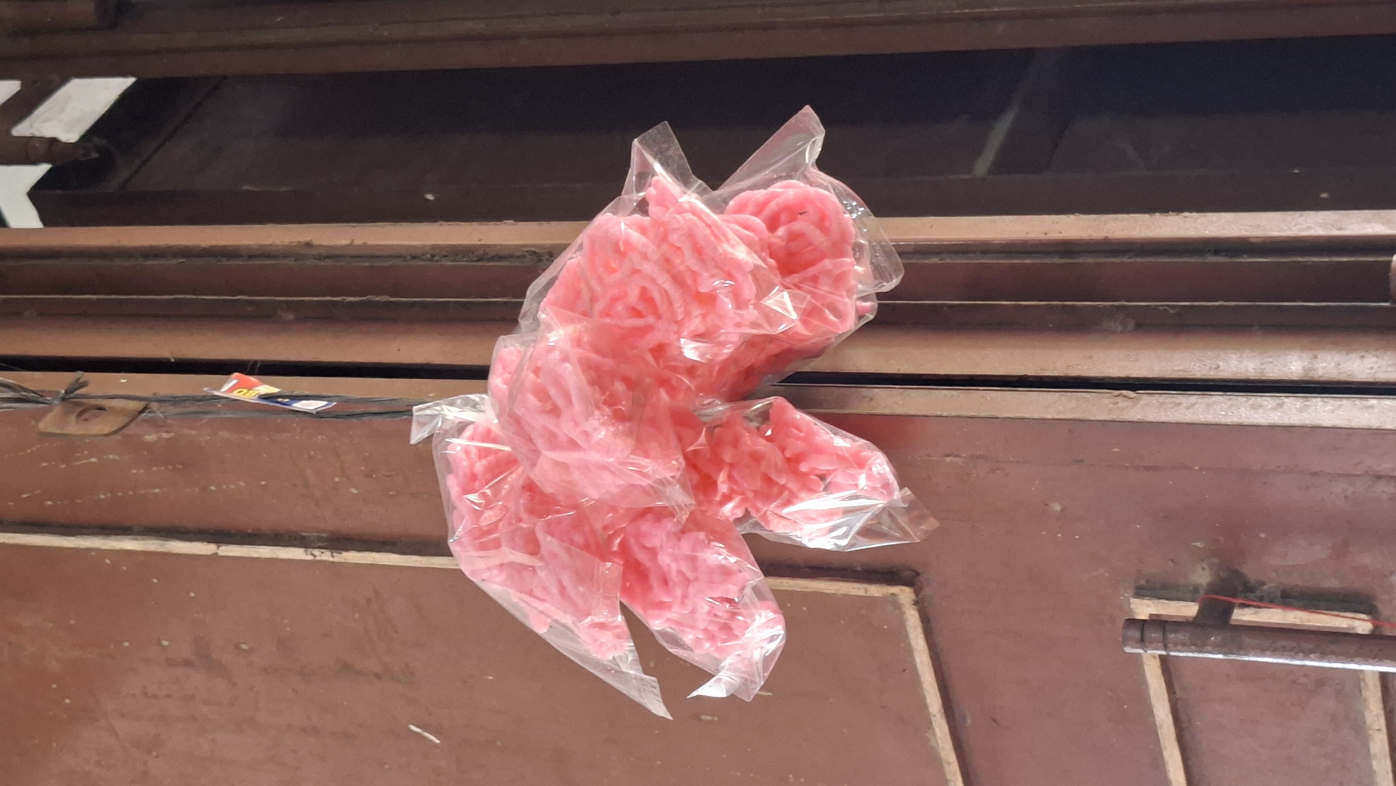 A bunch of pink crackers, possibly prawn crackers or krupuk, are hanging by a string against a weathered brown door. The crackers are individually wrapped in clear plastic, and several of these packs are clustered together. A small tag with red and blue colors is also attached to the string. The door itself appears to be made of metal and has vertical panels. A rusty metal pipe or railing is visible in the lower left corner of the image, attached to what looks like a wall or another part of the structure. The scene suggests an outdoor setting, perhaps a shop or stall in Keudebayu, Indonesia.