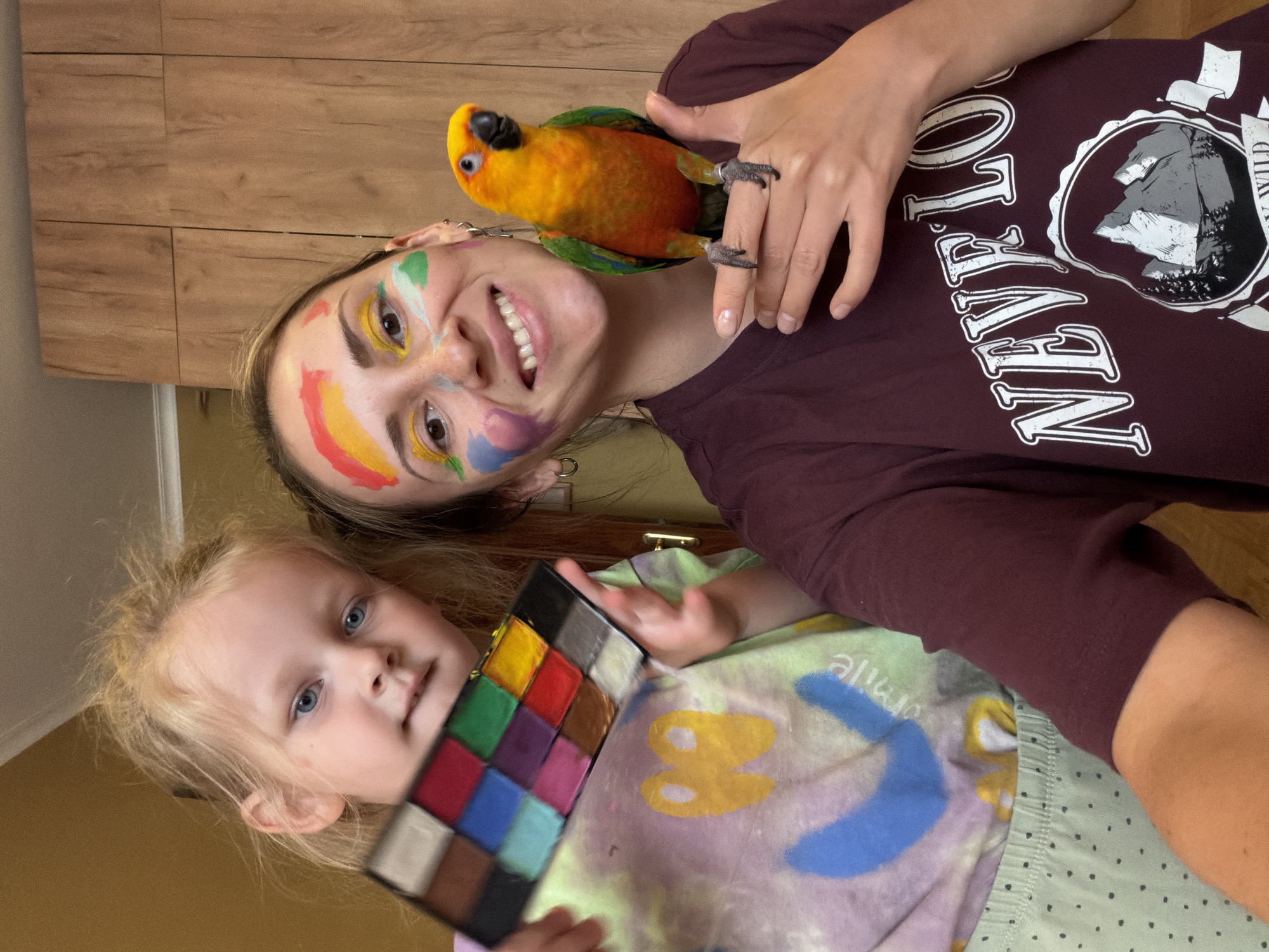A woman and a young girl are indoors. The woman is holding a parrot on her hand, smiling, and has face paint on her face, including a rainbow design. She is wearing a maroon t-shirt with a graphic design. The girl is holding a palette of face paints. The location is possibly Khimki, Russia.