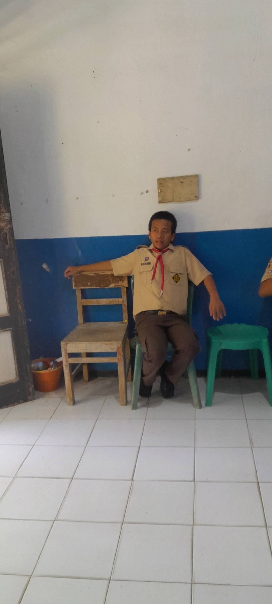 In Tahunan, Indonesia, a young boy in a scout uniform sits on a green plastic chair next to a wooden chair. He wears a tan shirt with a red scarf tied around his neck and brown pants. His left arm rests on the back of the wooden chair, and his right arm is extended. A brown bucket with a few items inside is on the floor to the left of the wooden chair. The room has white tiled flooring and walls painted blue and white.