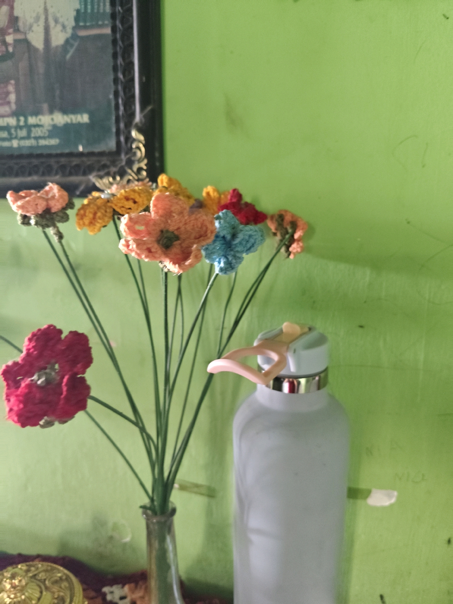 An overhead shot shows a bouquet of crocheted flowers, in shades of red, orange, yellow, and blue, arranged in a glass vase. The vase sits on a surface with a gold, ornate object to its left. Behind the flowers, a light blue water bottle with a silver rim and a pink carrying strap is visible. The background is a solid green wall. A framed photograph hangs on the wall above the flowers. The photograph displays a picture of a building with text that reads "MPN 2 MOJOKERTO" and "5 Juli 2005". The overall setting appears to be indoors, likely in a domestic or office space in Sumberwono, Indonesia.