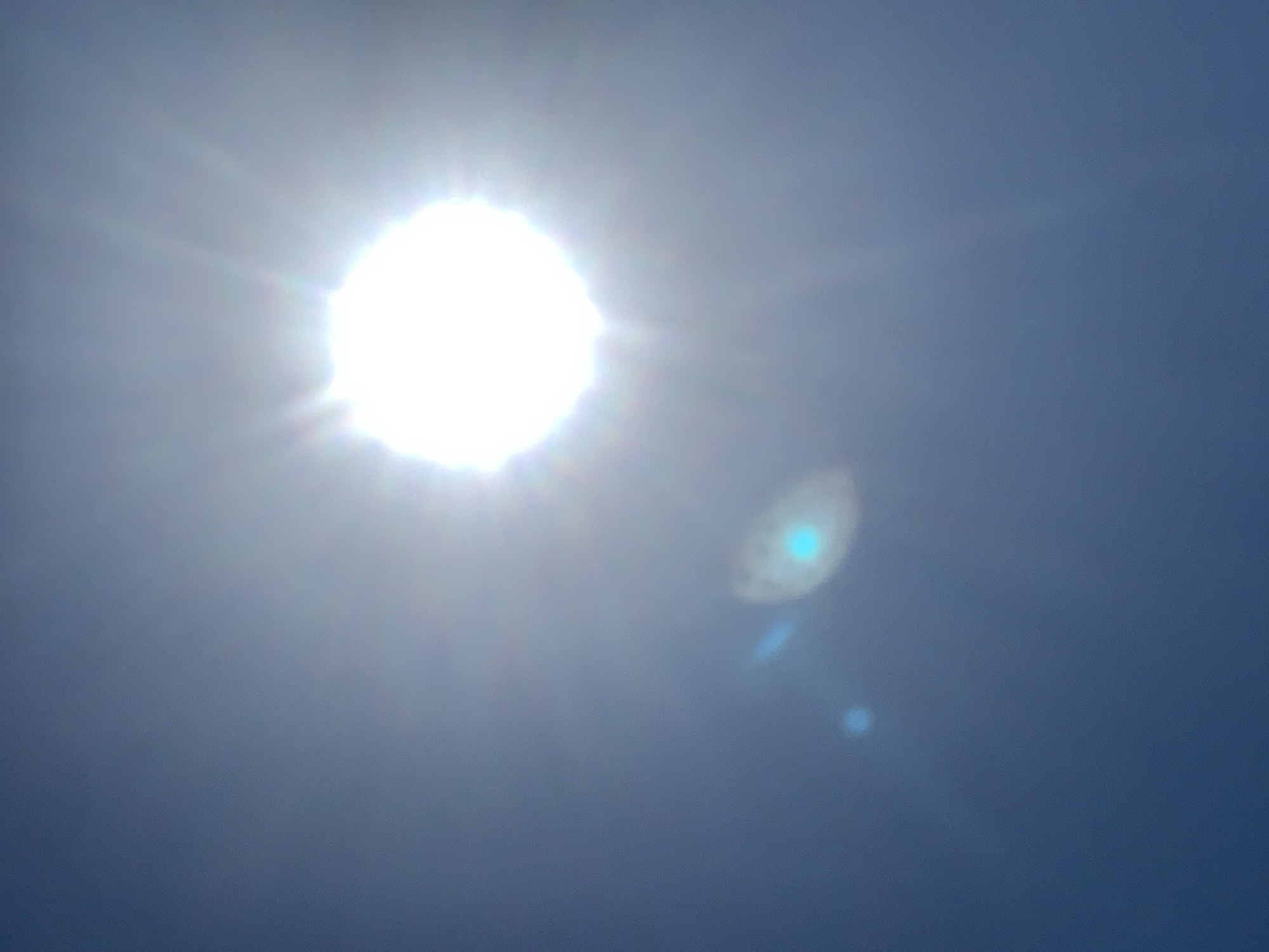 The image is an outdoor, close-up, full shot of the sun, taken during the daytime against a deep blue sky. The sun appears as a bright, white, circular object, with light rays emanating outwards. There are also several smaller, circular light flares present, one of which has a distinct blue hue, and another appears as a faint blue dot. The overall impression is that of a very bright day with the sun prominently featured, and the image is likely from Khazar Raion, Azerbaijan.