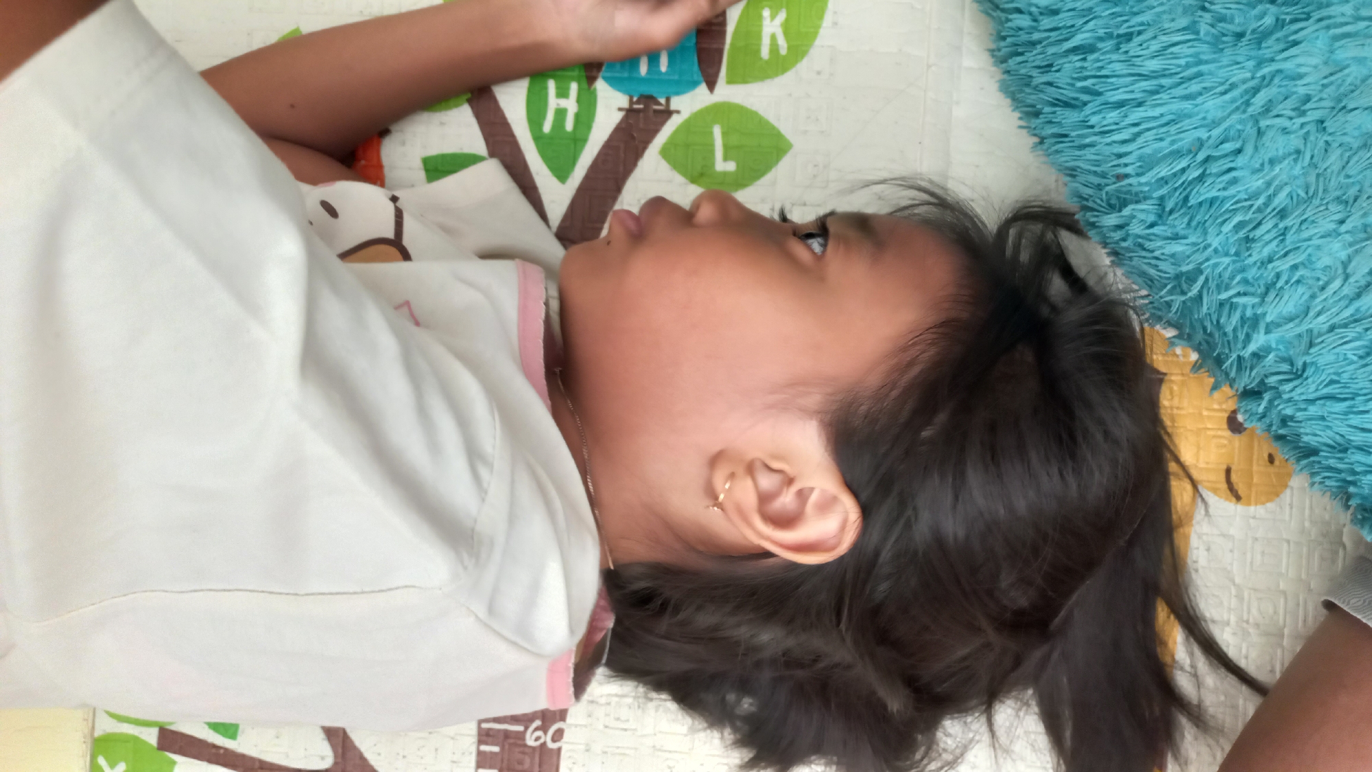 A young girl with dark hair is lying on her side on a playmat with animal prints. She is wearing a white t-shirt with a graphic of a bear. Her head is resting on a soft, teal-colored blanket. The playmat has green leaves with letters on them, including "H", "L", and "K". The image appears to be taken indoors, likely in a home setting. There are no visible indicators suggesting a specific location within Jetis Indah, Indonesia, beyond it being a domestic scene.