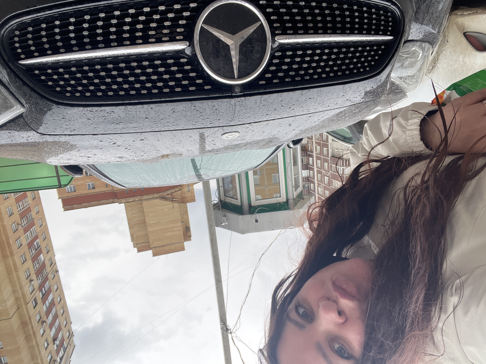 The image, when properly oriented, presents a close-up, selfie-style shot featuring a woman, a Mercedes-Benz car, and urban buildings under an overcast sky. The scene is located in Perm, Russia.
The dominant subject in the upper-left and central part of the frame is the front grille of a black Mercedes-Benz car. The grille features a hexagonal mesh pattern, with a prominent silver Mercedes-Benz star logo positioned centrally. Just below the star, a small camera lens is visible, likely for parking assistance or a 360-degree view system. The car's surface is covered in numerous small water droplets, indicating recent rain or high humidity.
To the right, partially visible, is a woman. Her dark, wavy hair frames her face, with her left eye and nose clearly looking towards the viewer. She is wearing a light-colored jacket, possibly white or beige, whose ruffled texture is noticeable on the sleeve. Her hand is visible towards the bottom right, holding an object that appears to have green and white packaging, though specific details or text are unidentifiable.
The background, primarily on the left side, consists of several multi-story urban buildings. One tall, light brown or beige apartment block with numerous windows is prominent, and another structure with brick-red elements is visible further back. A green awning or roof section is also noticeable on the far left. The sky is overcast, contributing to a muted daylight ambiance.
The image captures a casual moment in an urban setting, featuring the car's distinct branding and a partial view of the individual, set against the backdrop of city architecture in Perm, Russia. No additional readable text is visible in the scene.
