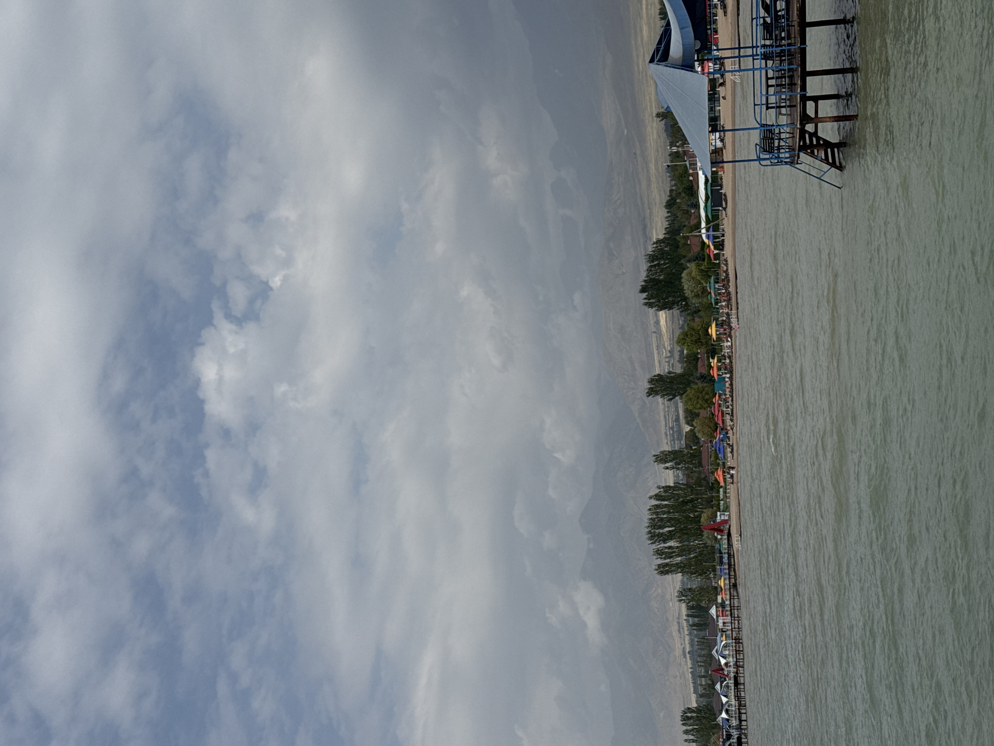 This image depicts a scene on the shore of Lake Issyk-Kul in Sary-Oy, Kyrgyzstan. In the foreground, a pier with a blue canopy extends over the water. The pier has seating and a ladder leading down to the lake. The water is a greenish-gray color and appears somewhat choppy. In the middle ground, a beach area is visible with numerous colorful umbrellas, lounge chairs, and small buildings or cabanas. A line of tall, slender trees runs along the edge of the beach. In the background, a range of mountains is shrouded in atmospheric haze and clouds. The sky is overcast with patches of lighter clouds. The overall impression is that of a resort area on a lake, with a backdrop of natural mountain scenery. There are no distinct people or specific actions clearly identifiable.