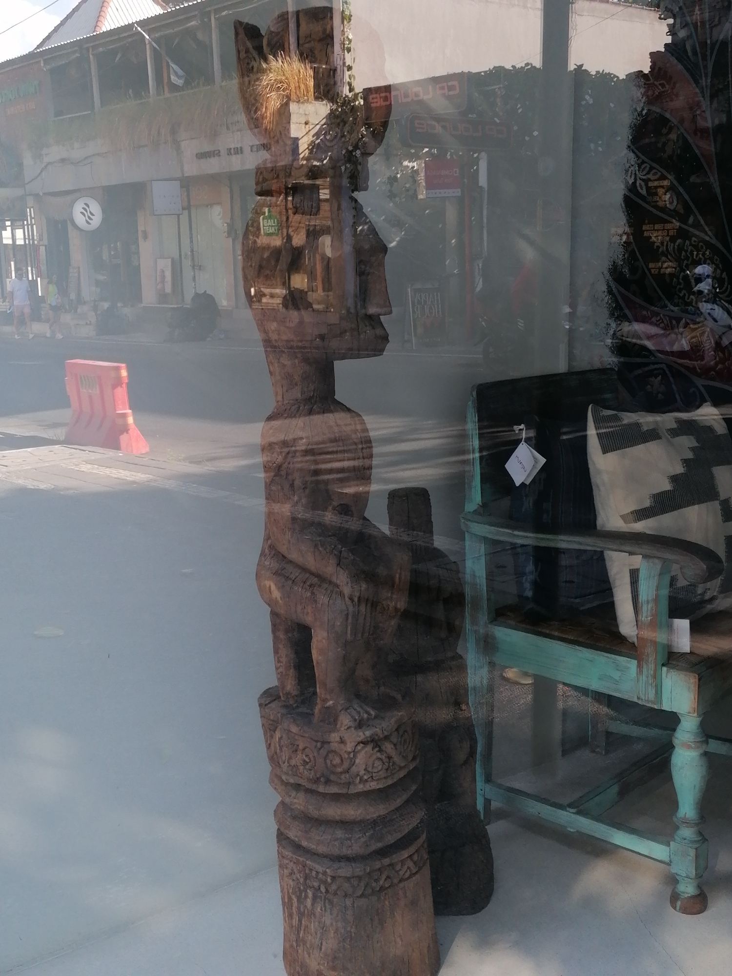 The image depicts a reflection in a storefront window in Ubud District, Indonesia. A carved wooden statue, possibly an Indonesian tribal art piece, is the primary subject in the foreground, reflecting from within the store. The statue is dark brown, with intricate carvings depicting a figure in a seated or crouching pose. It stands on a carved base.
Behind the statue, the reflection shows the interior of a shop, featuring a distressed teal-painted wooden bench with a cushion. The cushion has a black and white geometric pattern. A small white tag hangs from the bench.
The reflection also captures the street scene outside the shop. A red and orange plastic barrier is visible on the asphalt road. A building with a multi-story facade, possibly shops or restaurants, is seen in the background. The architecture appears to be typical of the region, with some signs and architectural details visible, including what looks like a circular logo with concentric rings on one of the storefronts. Sunlight casts shadows, and the overall scene is slightly blurred due to the reflection. No people are clearly identifiable within the reflection, but some figures are distantly visible on a sidewalk in the background.