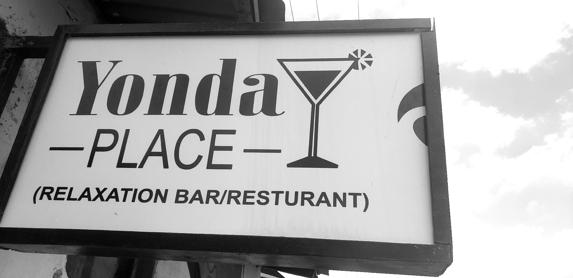 This is a black and white photograph of a sign for "Yonda Place - Relaxarion Bar/Resturant". The sign is rectangular with a dark frame. The text is in bold, dark lettering against a white background. The word "Yonda" is written in a serif font, and below it, "-PLACE-" is in a sans-serif font. To the right of the text is a stylized graphic of a martini glass with a lemon wedge. On the far right of the sign, there is a dark curved shape, possibly part of a logo. Below "PLACE-", in smaller sans-serif font, is the text "(RELAXATION BAR/RESTAURANT)". The sign is mounted outdoors, with a cloudy sky visible in the background, suggesting it is daytime. The overall impression is of a commercial establishment, likely a bar or restaurant, located in Narayi, Nigeria.