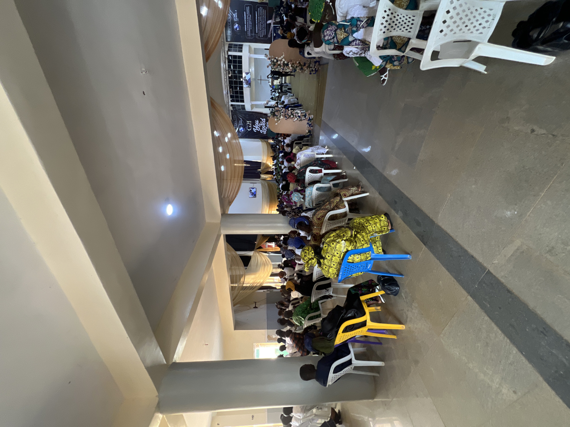 This image depicts an indoor gathering, likely a religious service or event, held in Kabong, Nigeria. The scene is set in a spacious hall with a high ceiling, featuring prominent beige beams and columns. A large crowd of people, predominantly women dressed in colorful traditional attire, are seated on rows of white and blue plastic chairs. The attendees are facing towards the front of the hall, where decorations, including floral arrangements and banners, are visible. Some banners display text such as "WELCOME" and "ECWA GOSPEL 2". The floor is tiled in large, muted-colored stone slabs, with a dark grey strip running diagonally across the foreground. Lighting appears to be a mix of natural light from windows and artificial ceiling lights, creating a warm ambiance. The overall atmosphere suggests a community event in progress.