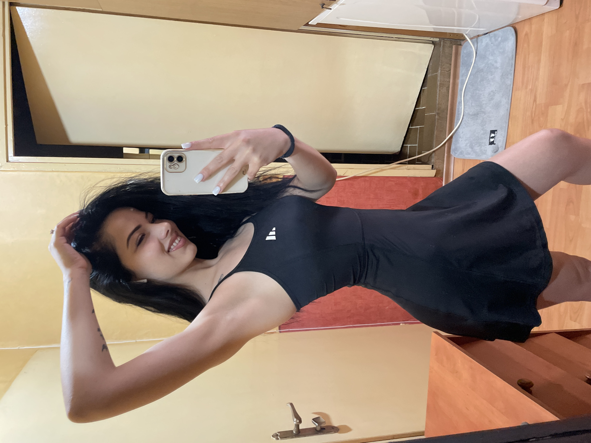A young woman with long black hair and fair skin takes a selfie in a room with light yellow walls and a wooden floor. She is wearing a black sleeveless athletic dress with a white Adidas logo on the chest and white painted fingernails. Her right arm is raised and her hand is in her hair, and she is smiling. She is holding a white smartphone in her left hand, which is capturing the reflection of herself and the room in a mirror. To the left of the woman is a wooden dresser. To her right is a closed door with a metallic handle, and beyond that, a floor mat and a white appliance. The overall impression is that of a casual, personal moment captured within a domestic setting. No specific landmarks or indications of Stara Zagora, Bulgaria, are visible in the image.